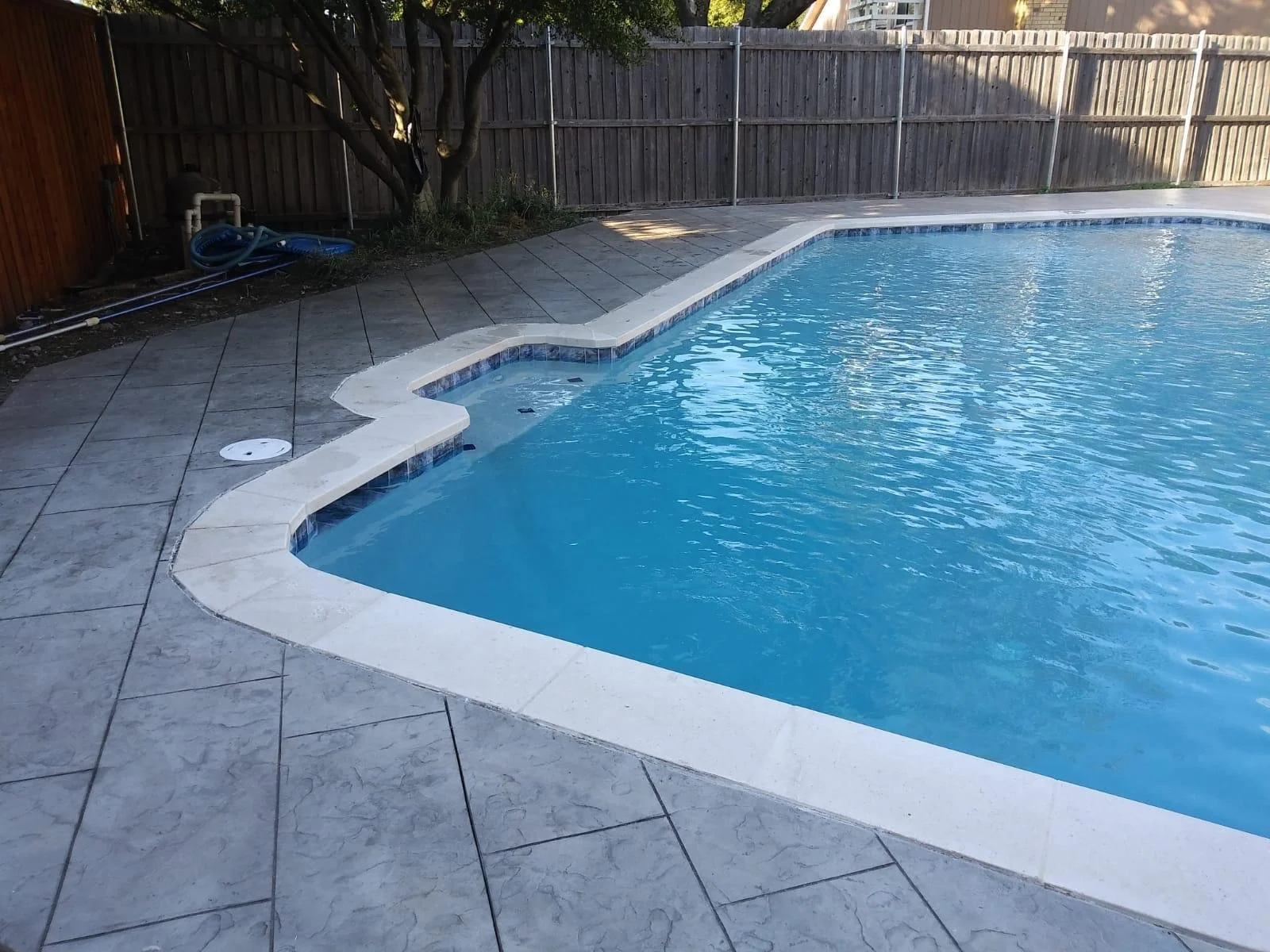 Residential backyard swimming pool with gray tiled deck and wooden fence, with some pool equipment and hose near a tree.