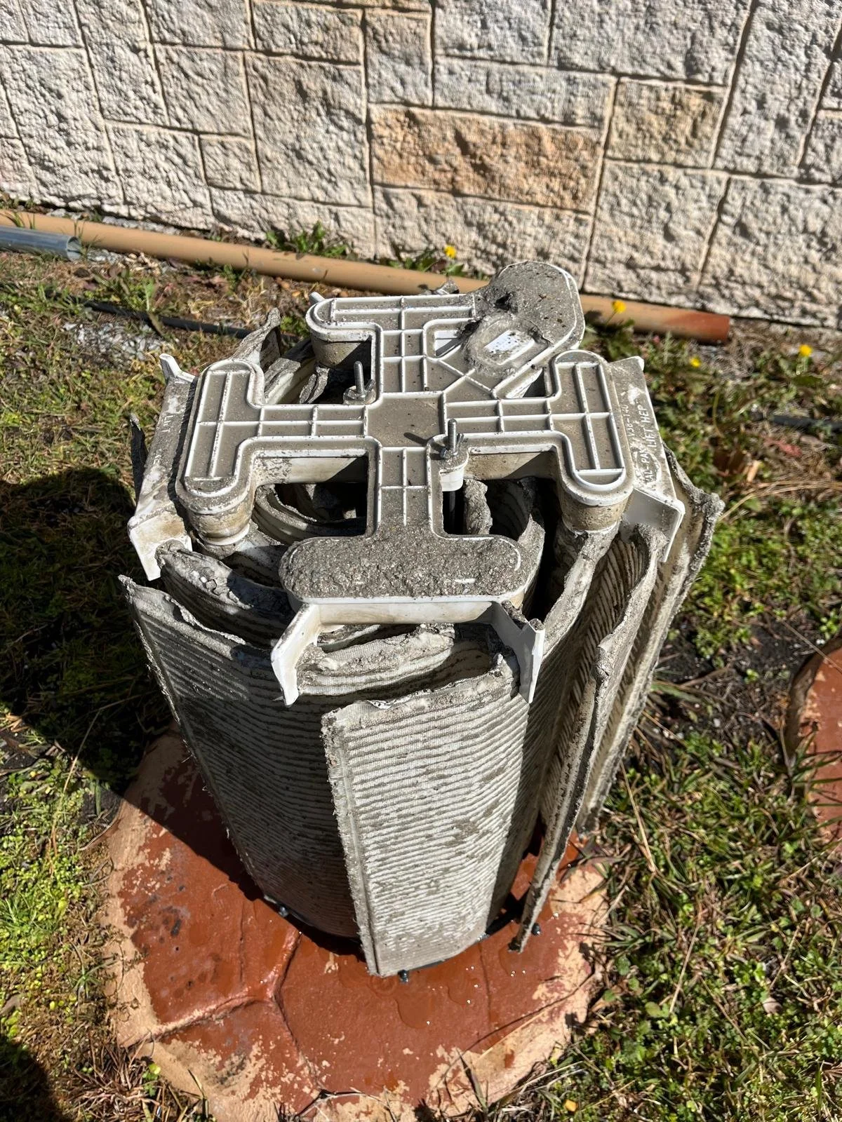 A damaged air conditioning condenser unit outdoors with visible bent metal fins and debris on top, sitting on a concrete slab next to grass and a brick wall.