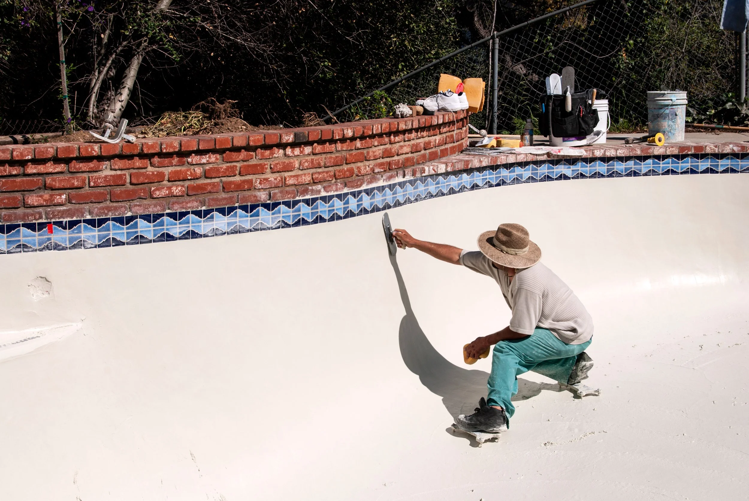 A person working on a pool's tile border, using a tool, wearing a wide-brimmed hat, and surrounded by various tools and supplies, with a brick wall, fence, and plants in the background.