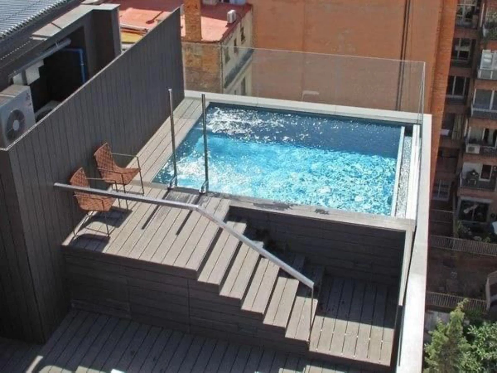 Rooftop swimming pool with wooden deck lounging area, two brown chairs, metal railing, and city buildings in the background.