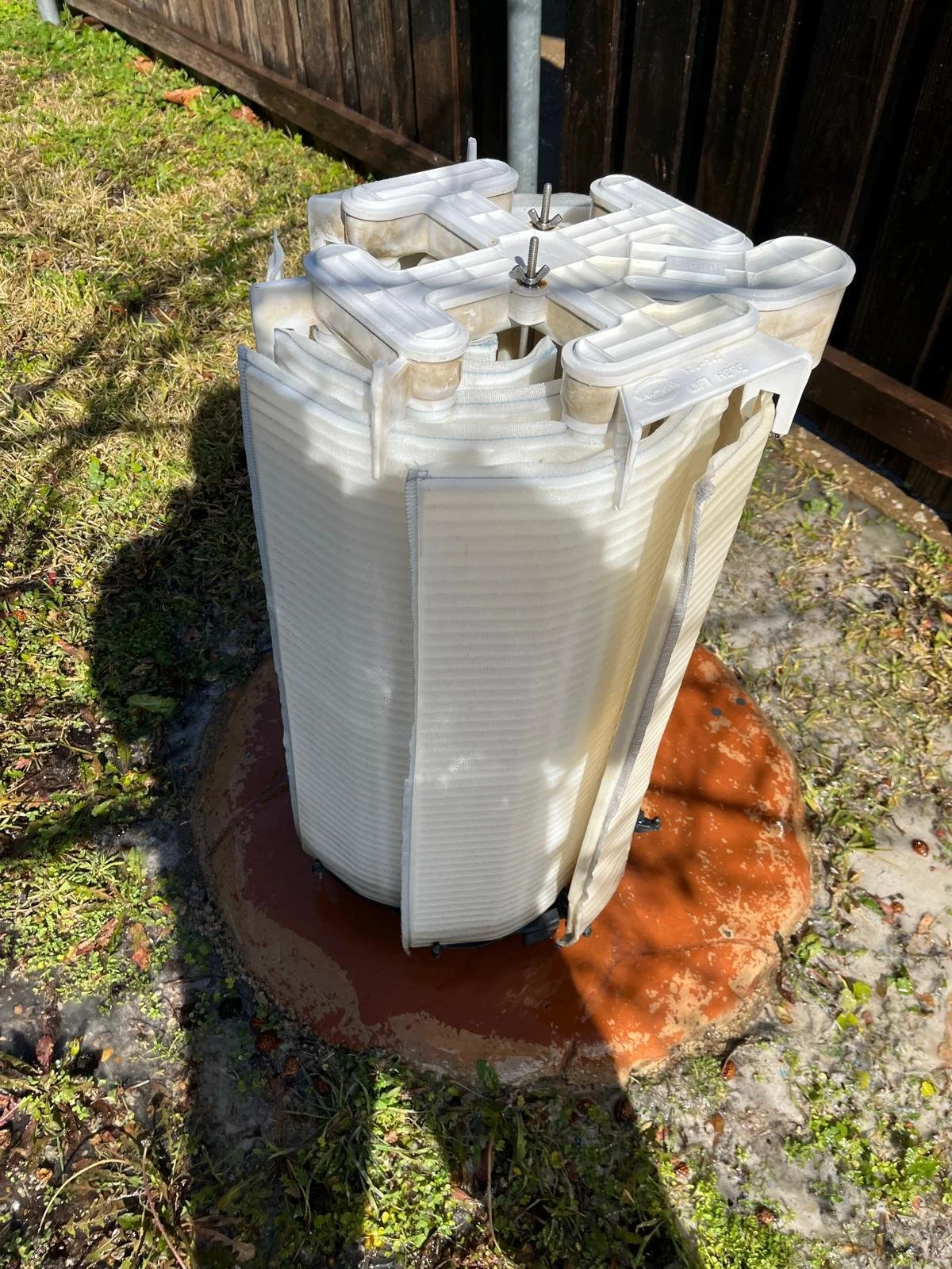 A white, cylindrical, water softener or filtration system with tubing and bolts on top, installed outside on a concrete pad, next to a wooden fence and surrounded by grass and small plants.