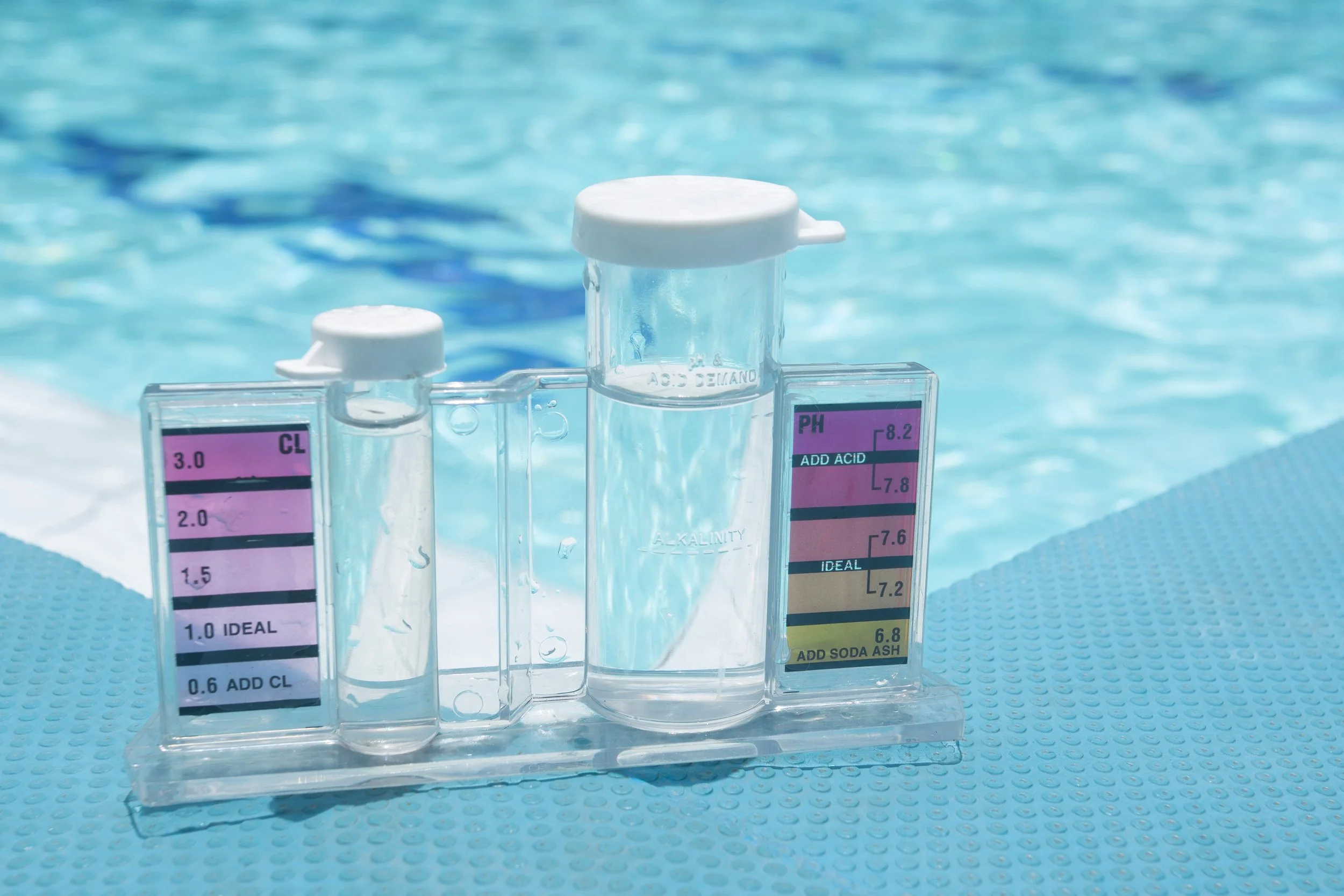 A set of water testing vials with color-coded labels, placed on a light blue textured surface, with a swimming pool in the background.