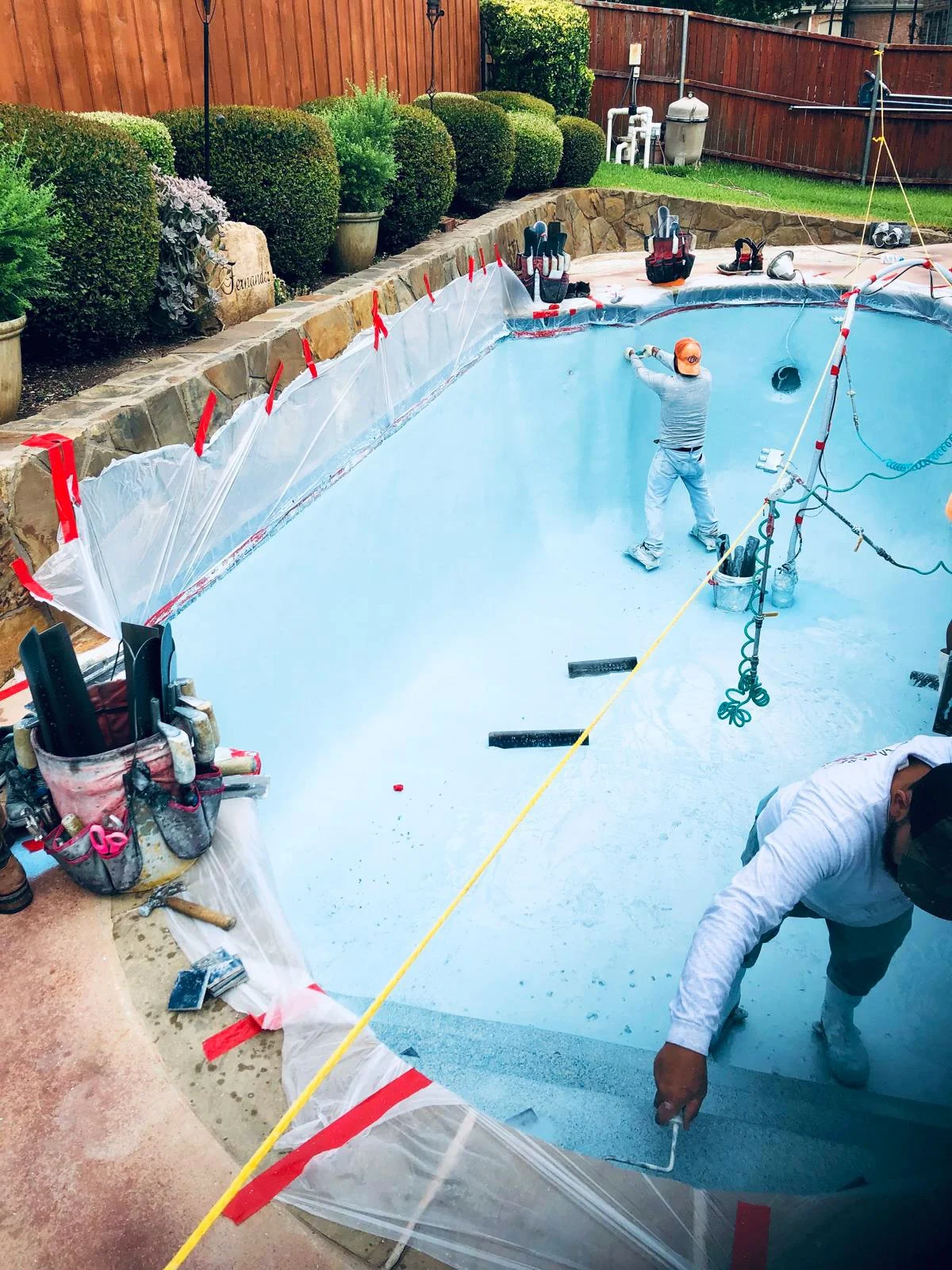 Workers painting the inside of a backyard swimming pool with blue waterproof paint. The pool is surrounded by a stone border, and construction tools and equipment are visible nearby. There is a wooden fence and landscaped bushes in the background.