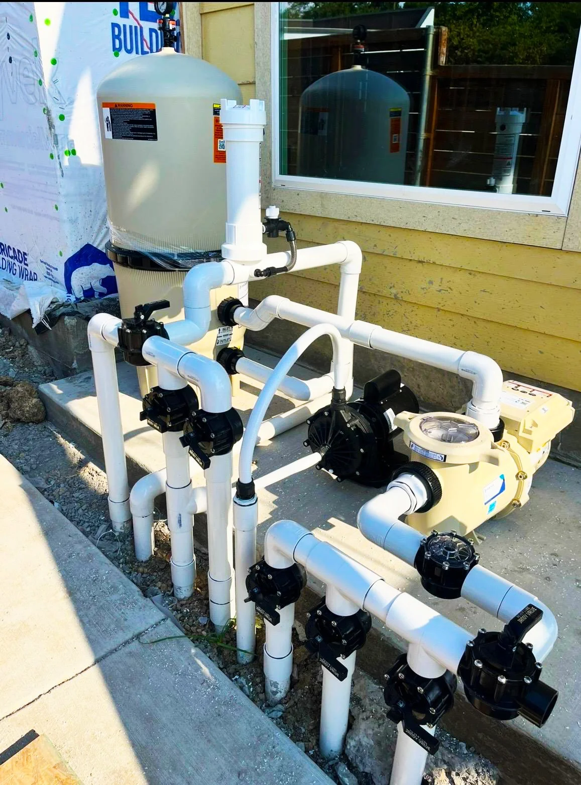 A pool filtration system with white PVC pipes, black valves, a sand filter, and a pool pump installed outside next to a yellow house wall with a window.