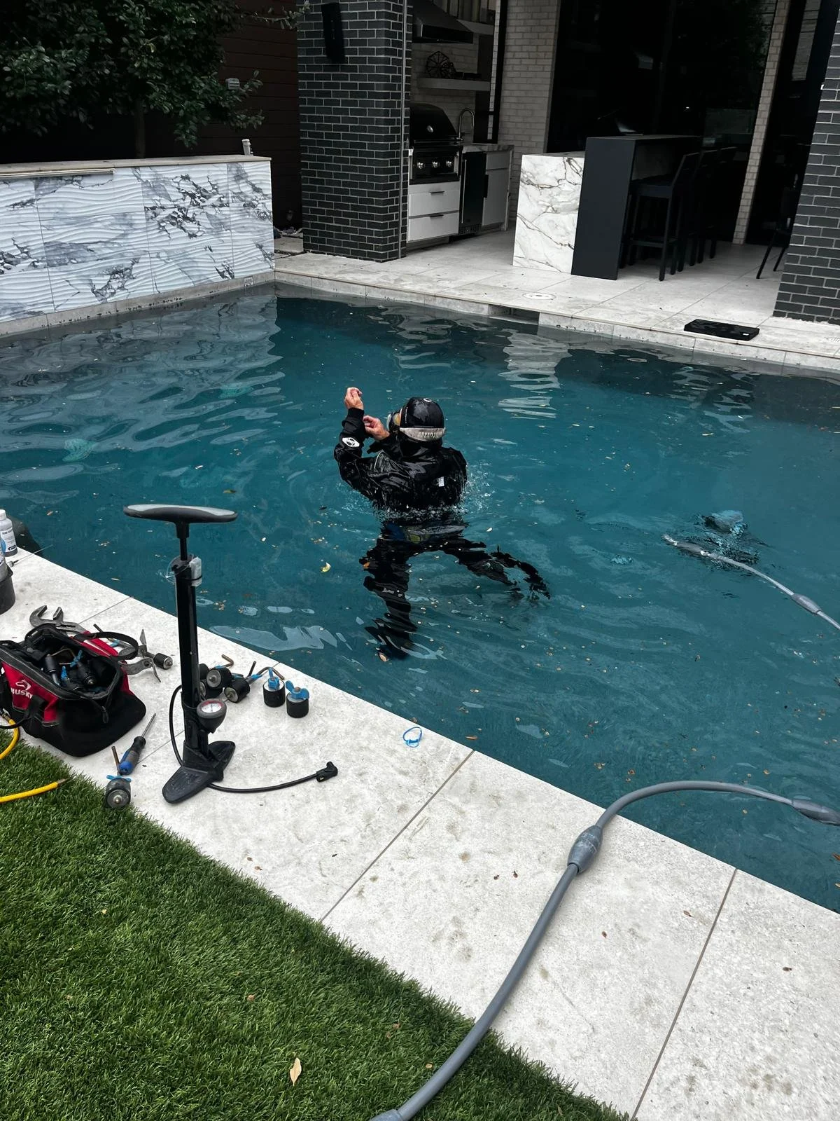 Person in black wetsuit and diving gear working in a swimming pool, surrounded by tools and equipment.