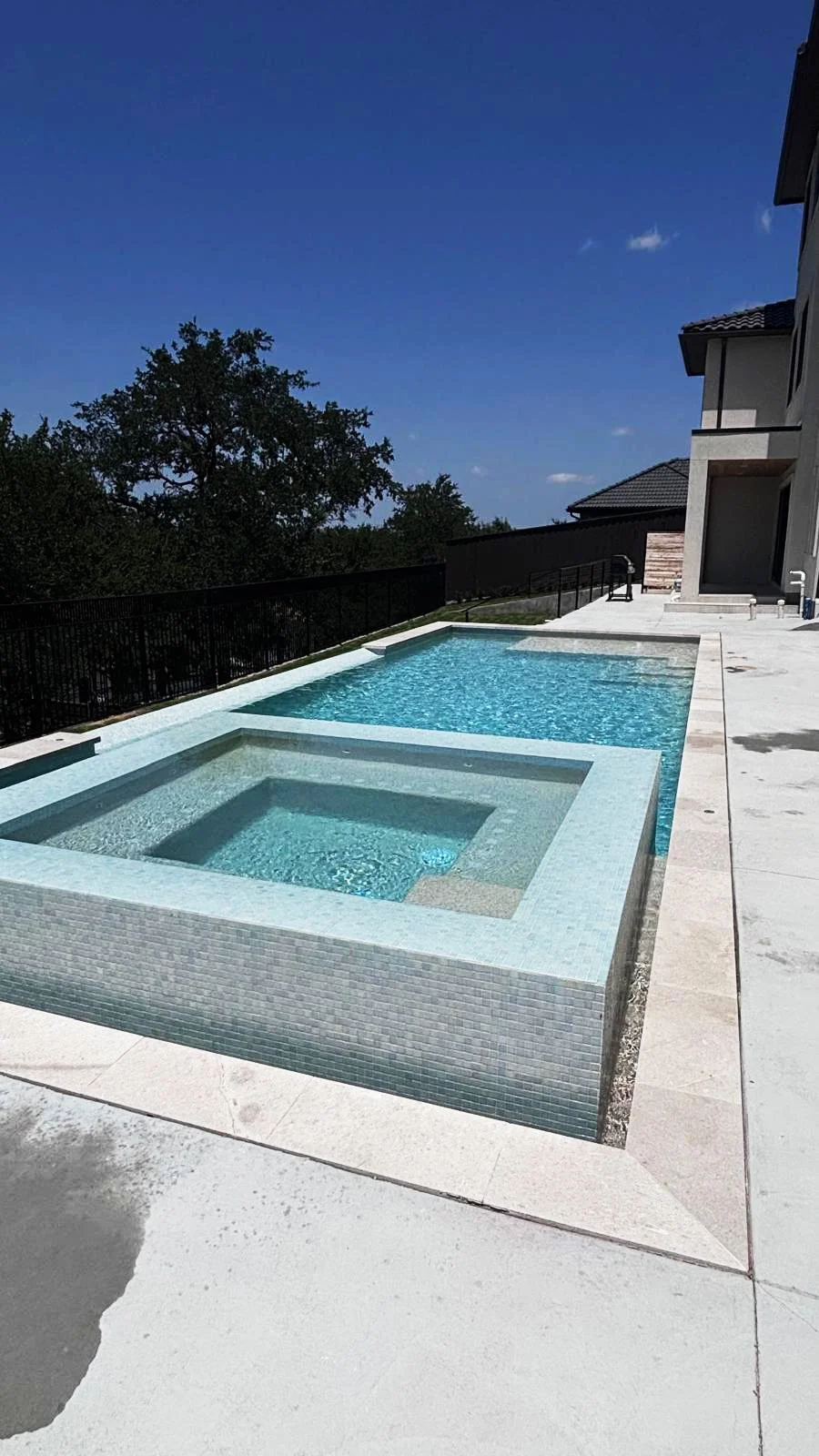 View of a modern backyard swimming pool with an attached hot tub, surrounded by concrete patio, with a black metal fence, and a house with balconies in the background under a clear blue sky.