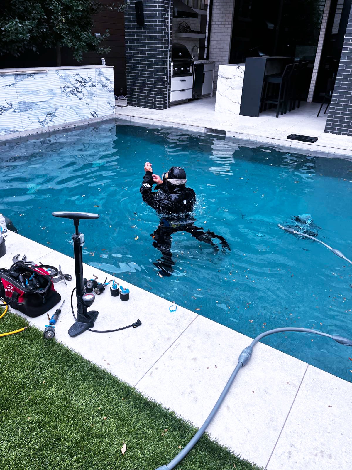 A person in black diving gear and a helmet is in a backyard swimming pool, performing maintenance with tools on the poolside. There is equipment and tools such as a vacuum, hoses, and a toolbox nearby, with a charcoal grill and outdoor kitchen area in the background.