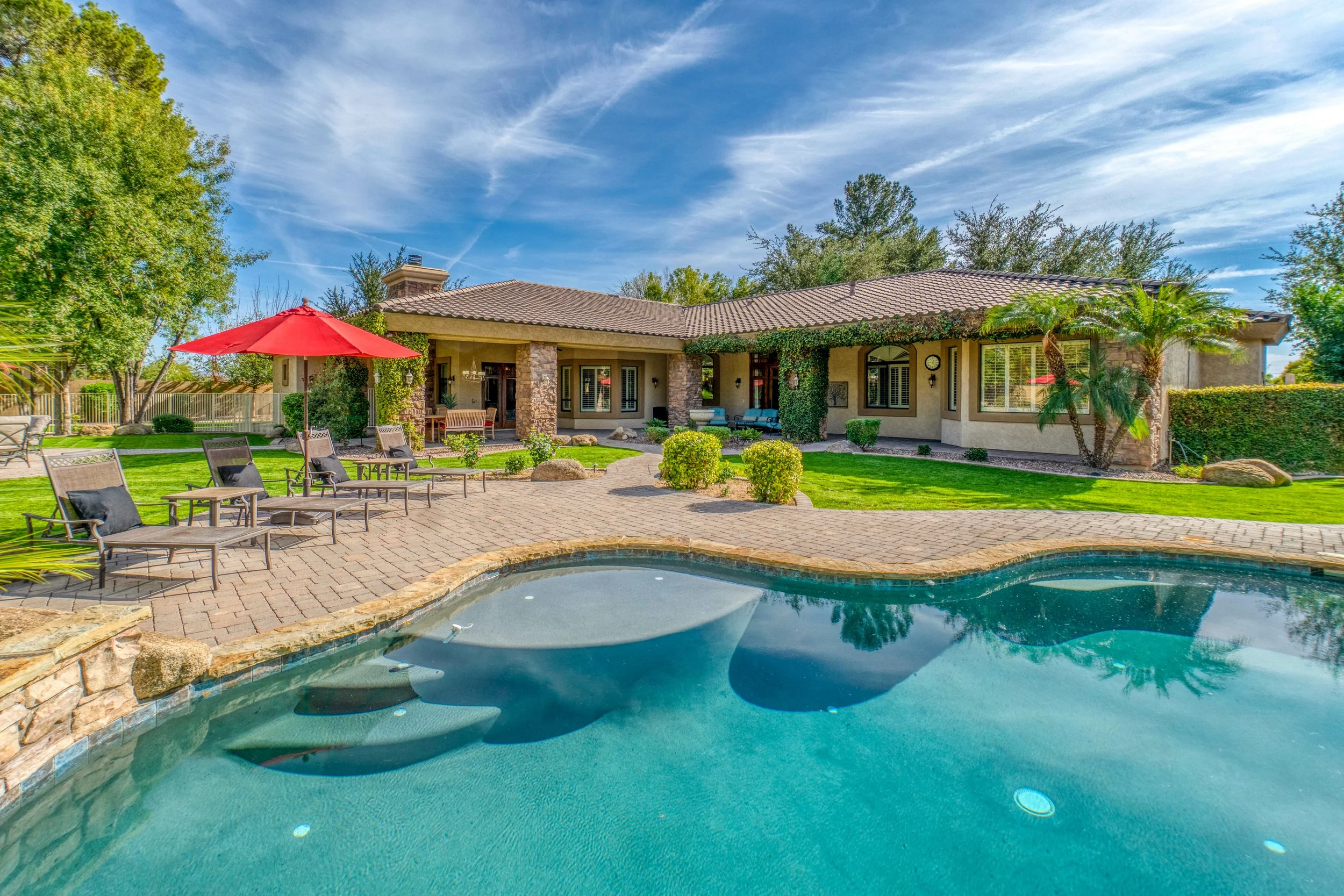 Backyard with swimming pool, patio furniture, a red umbrella, and a house with trees and a blue sky in the background.