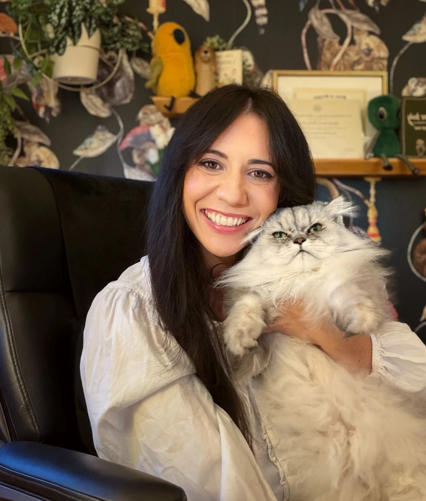 A woman with long dark hair smiling and holding a fluffy gray cat. The background features a black wall with decorative art and framed pictures, including a yellow plush bird and various plants.