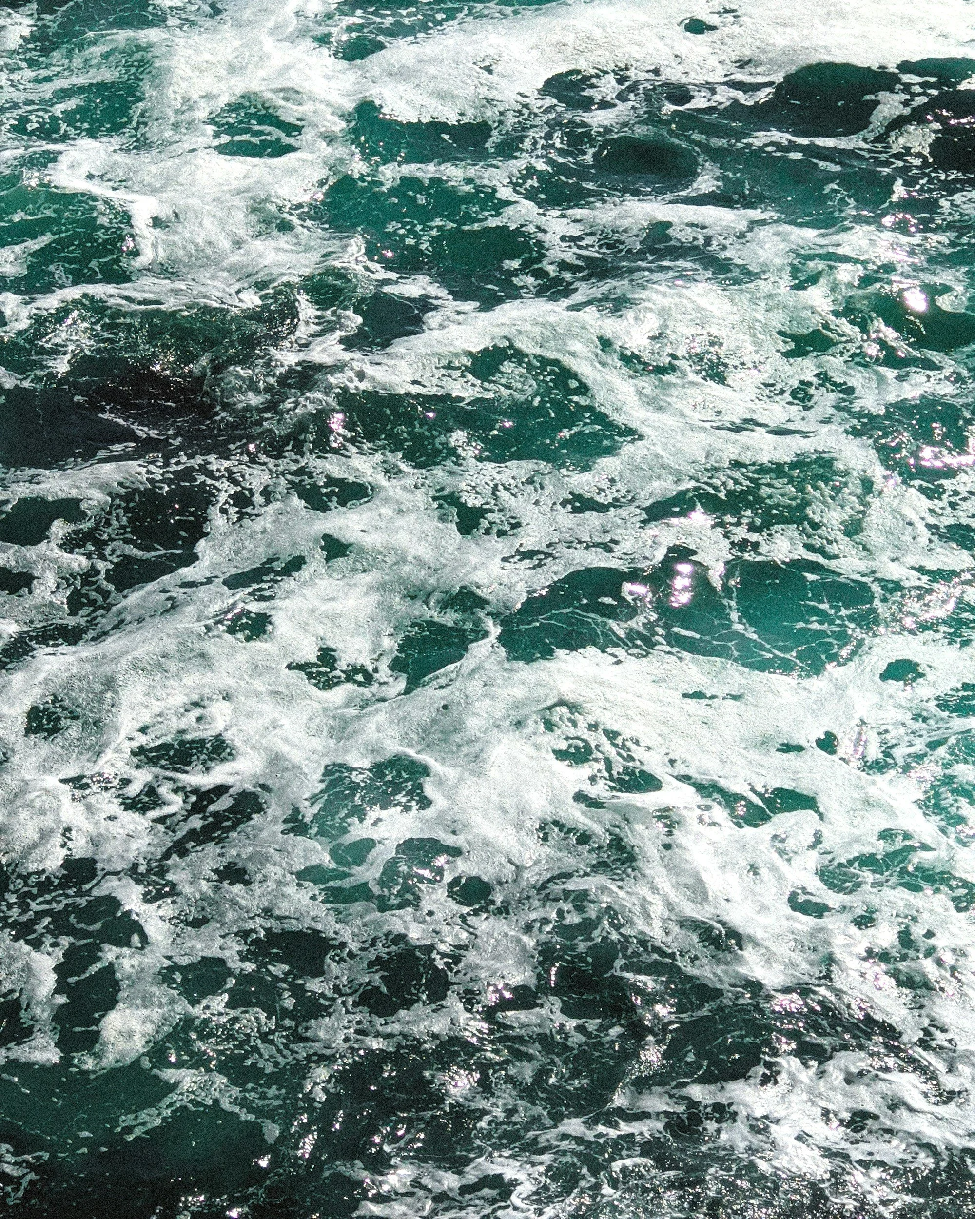 Close-up view of ocean water with white foamy waves and surf, sunlight reflecting off the surface.
