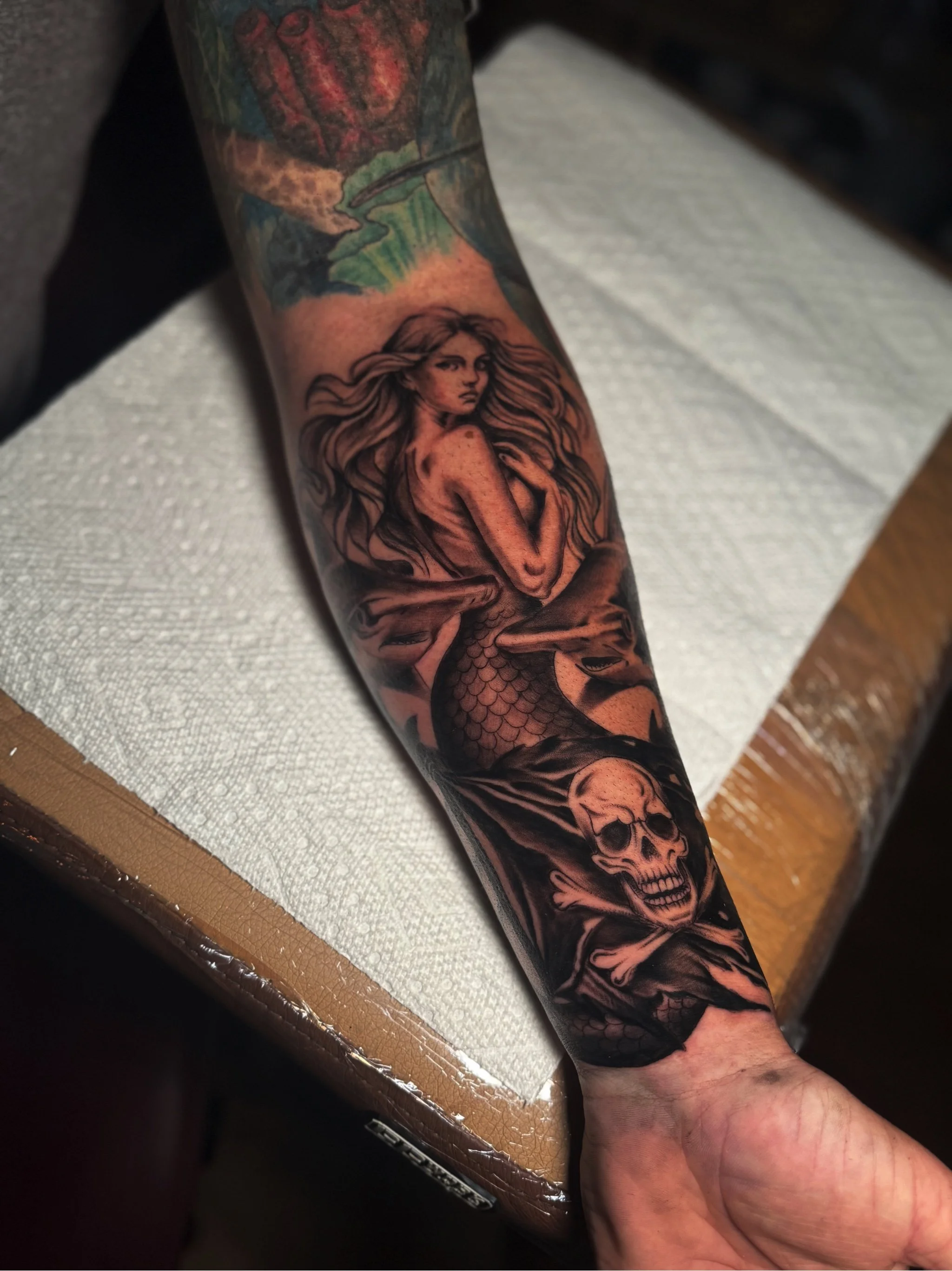 Tattoo of a woman with long flowing hair on the forearm, with a detailed skull tattoo below it, and background elements including a bear's face and a landscape scene with trees.