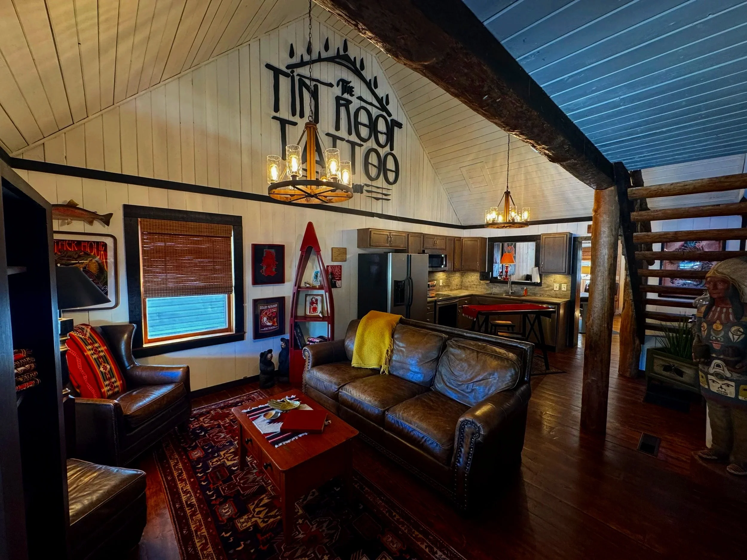 A cozy living area with leather sofas, a patterned rug, framed wall art, and wooden ceilings with chandeliers, in a cabin-style interior.