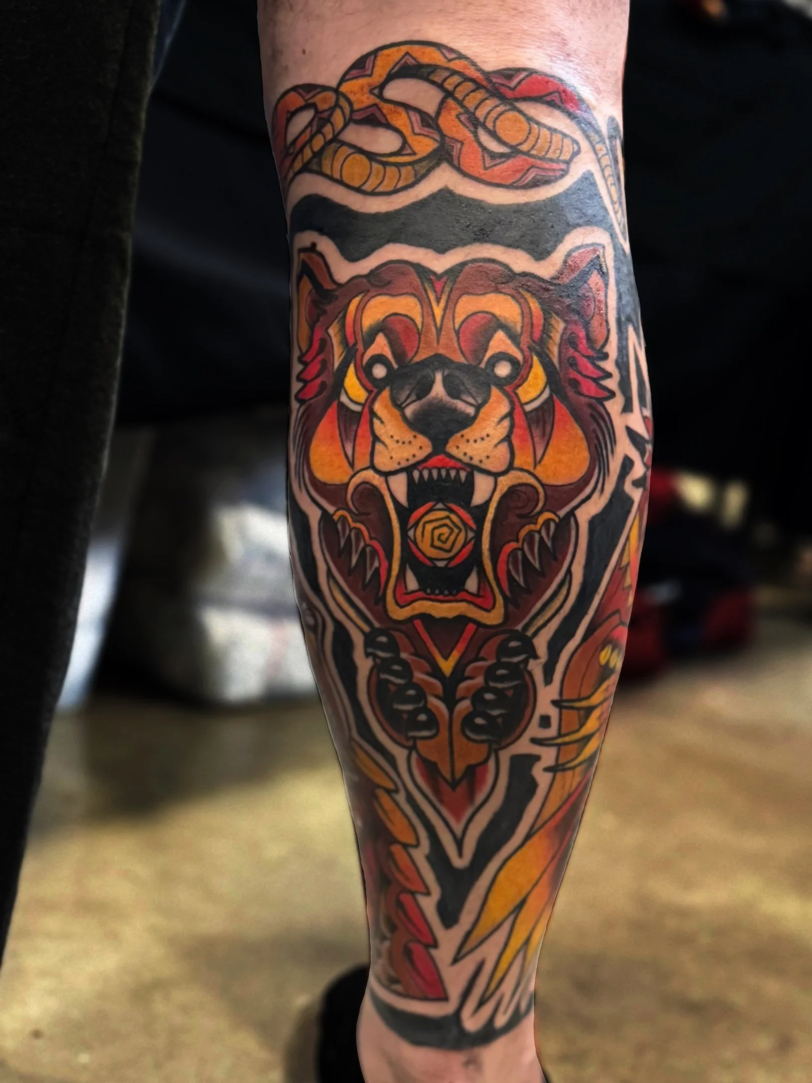 Colorful tattoo of a fierce lion's face with bold lines and vibrant orange, red, yellow, and black colors on a person's lower leg.