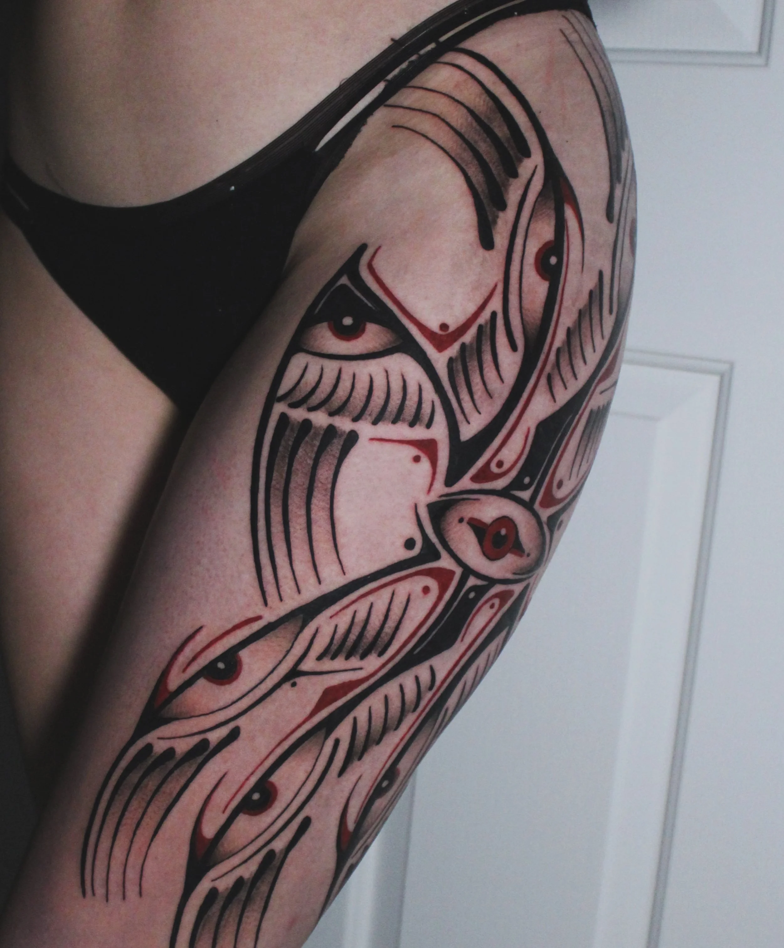 Close-up of a person's upper arm and shoulder with a tattoo. The tattoo features abstract black and red lines and eye-like designs. The person is wearing a black tank top. The background includes a white door with panels.