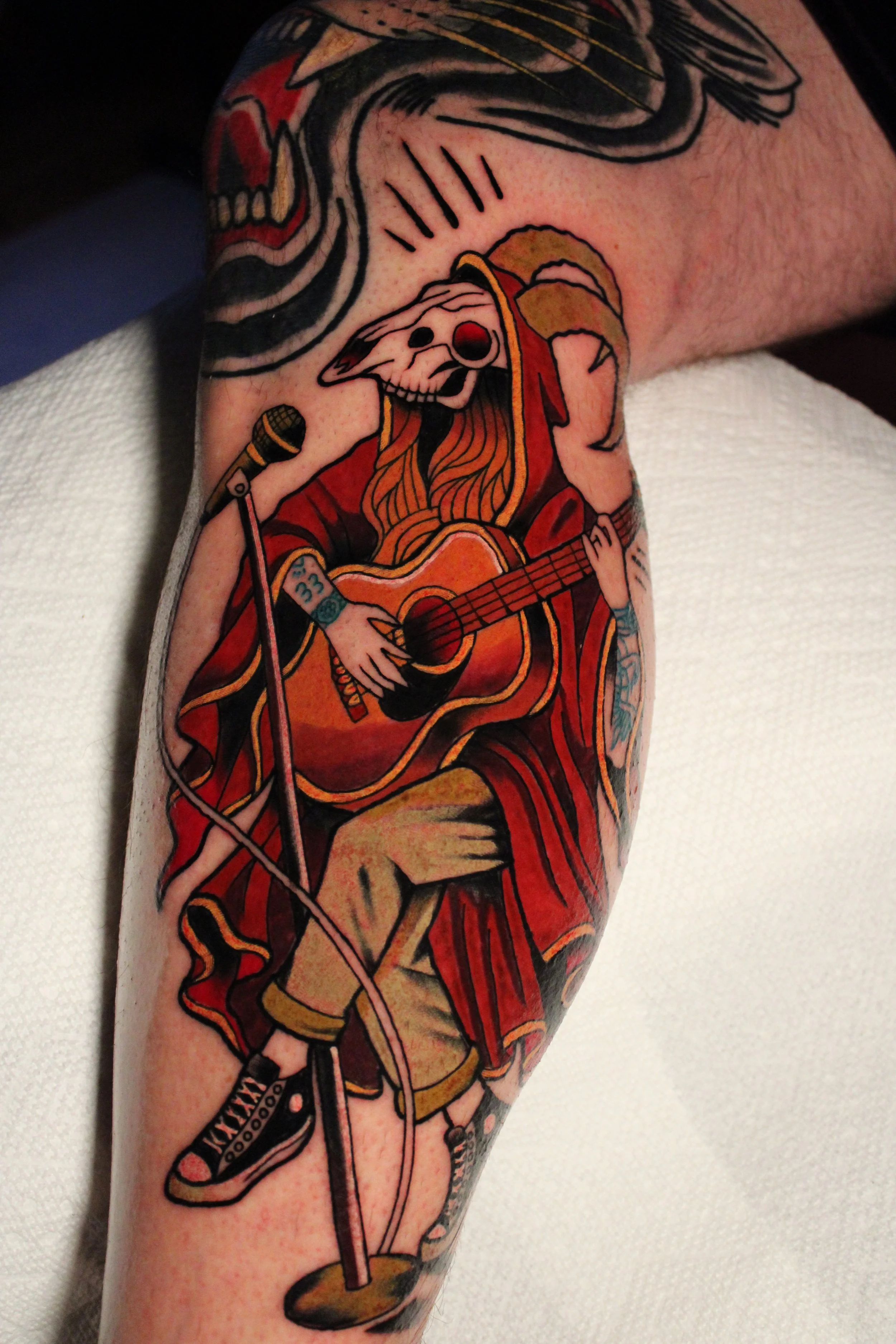 Tattoo of a skeleton playing guitar, singing into a microphone, wearing a red robe, with a large skull and bones, on a person's arm.