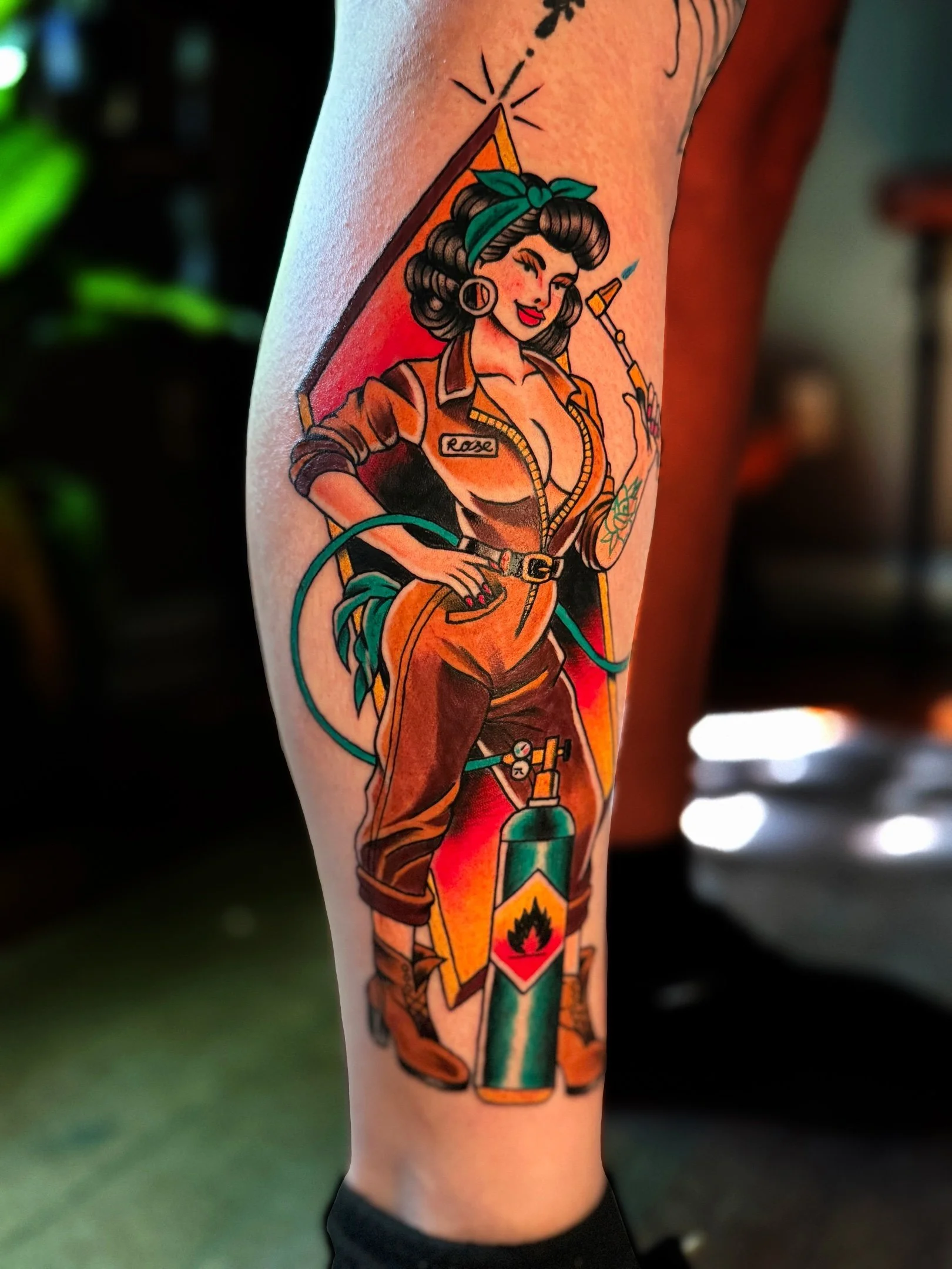 Tattoo of a vintage pin-up girl in a medical-themed outfit, holding a syringe and standing beside a torch with a flame, with the name "Rose" on her shirt.