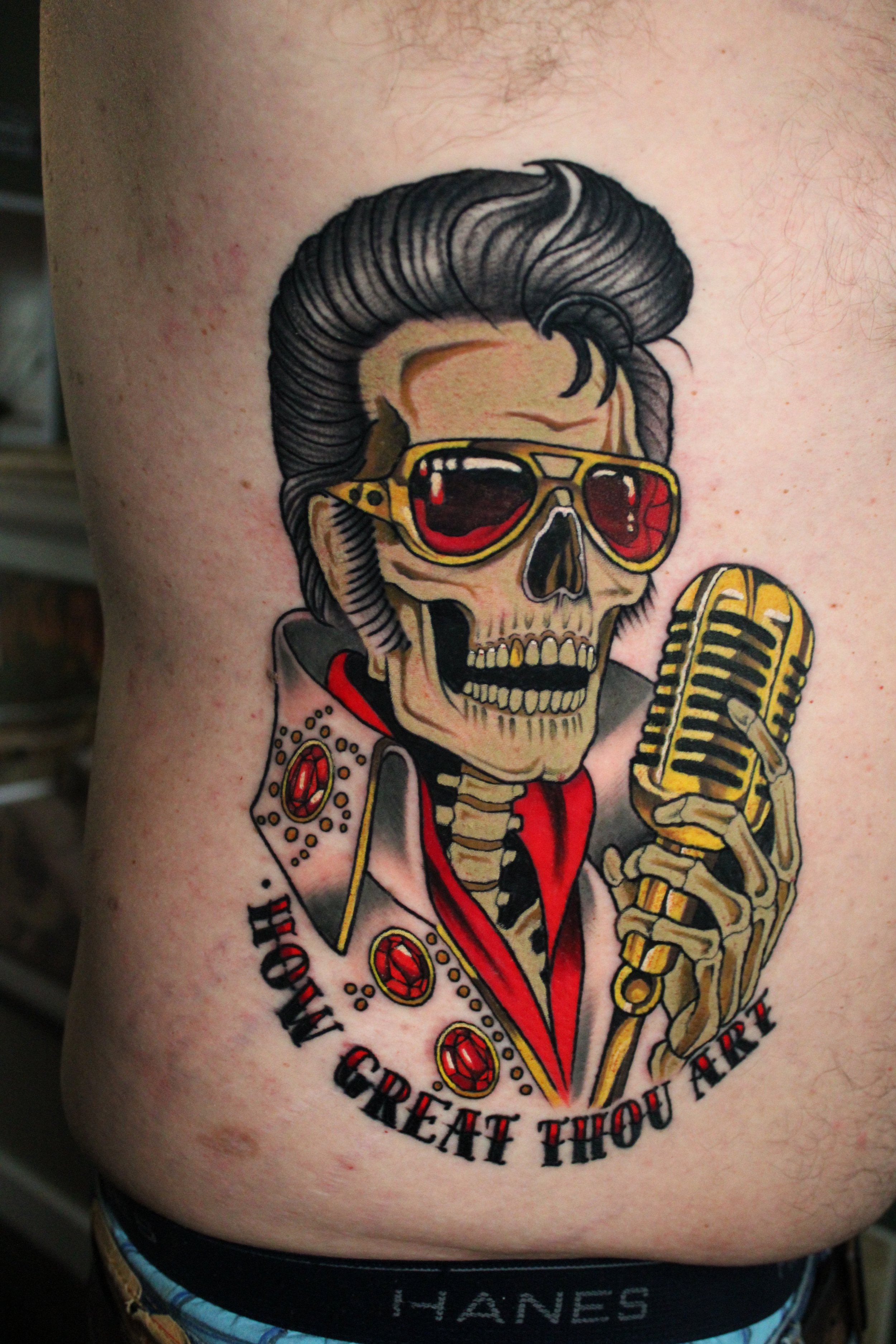 Tattoo of a skeleton in a red jacket with a skull wearing sunglasses, holding a vintage microphone, with surrounding text that says 'GREAT THOU ART'.