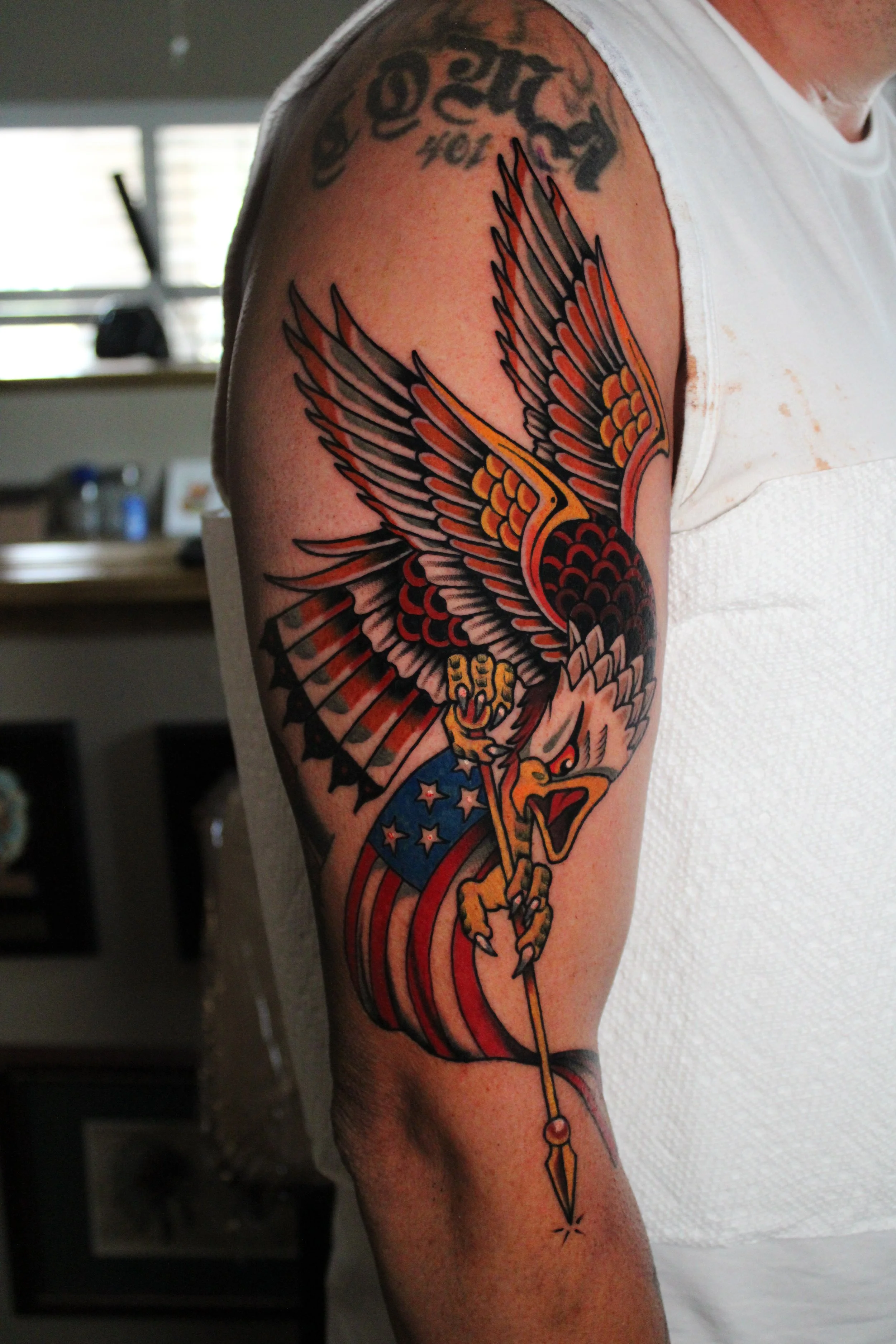 A person with a large colorful tattoo of a stylized eagle with outstretched wings, holding a sword, on their upper arm.