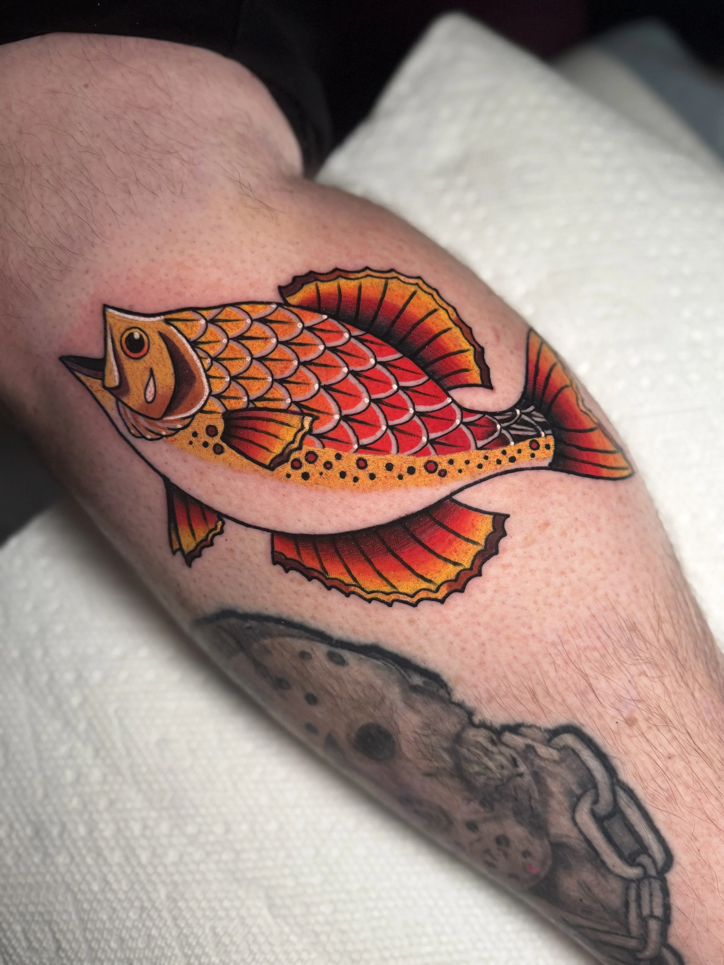 Tattoo of a colorful fish with orange, red, and yellow scales and detailed fins, on a person's arm.