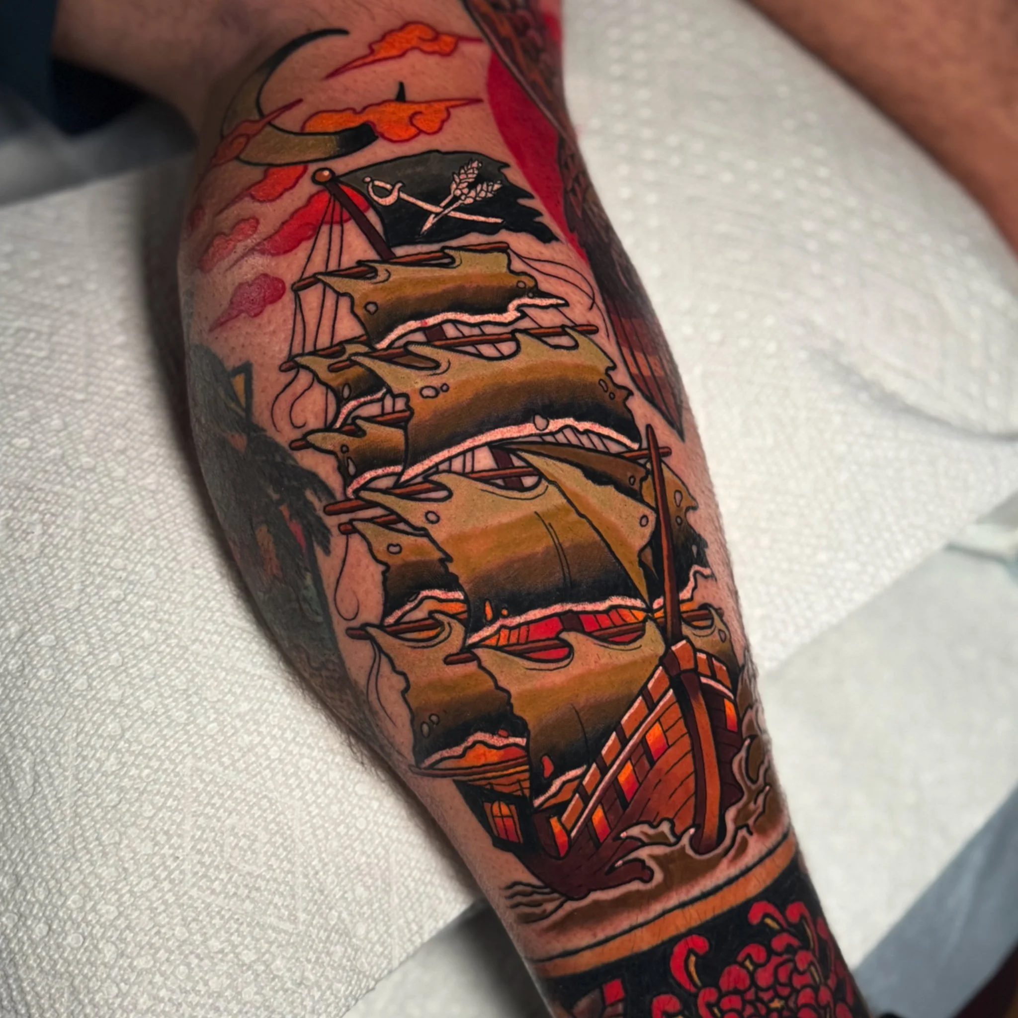 A tattoo of a pirate ship with black sails, an anchor, and a flag featuring a skull and crossbones, surrounded by orange and red flames on a person's arm.