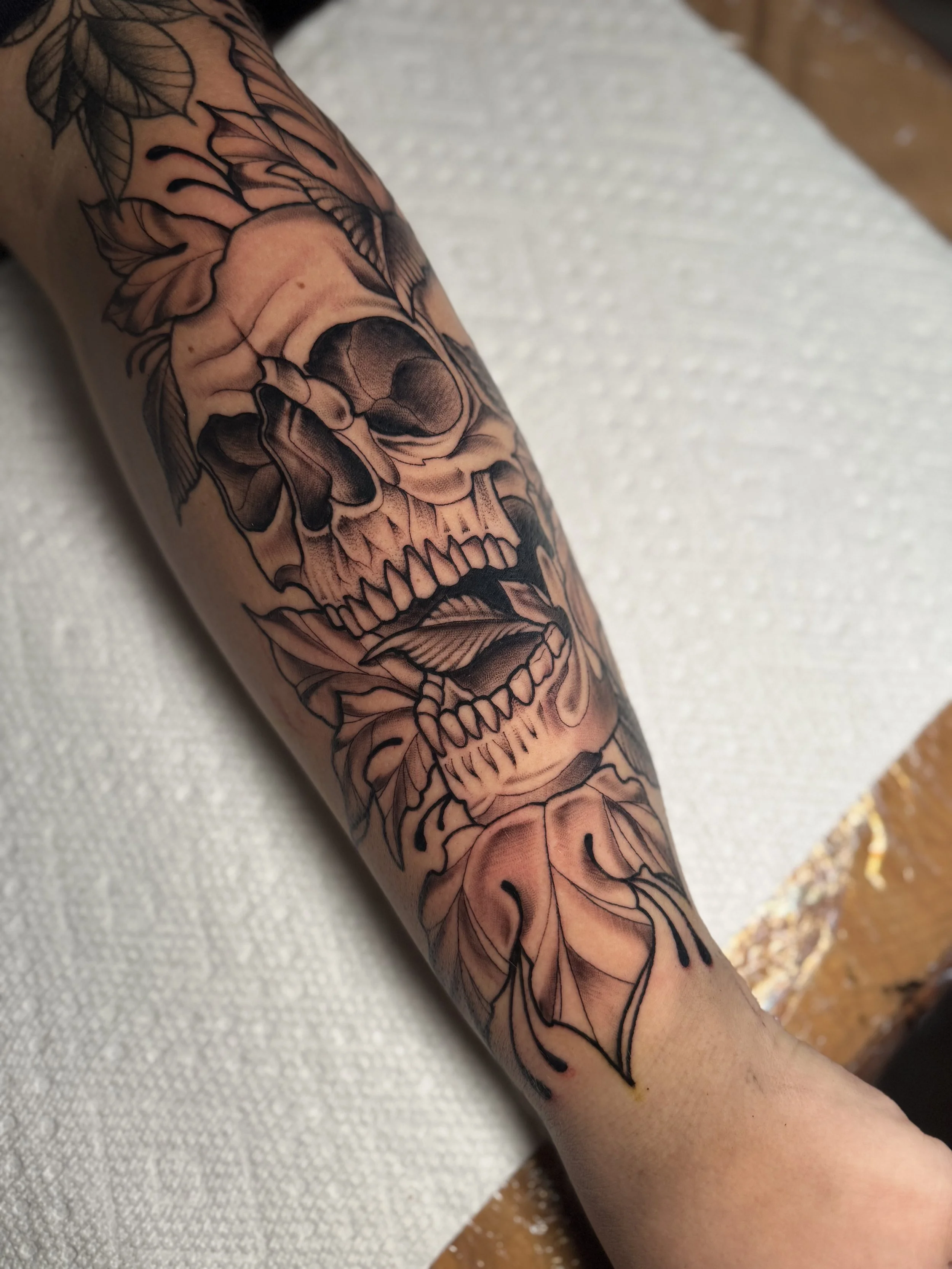 A person's arm with a black and gray tattoo of a skull with roses and leaves.