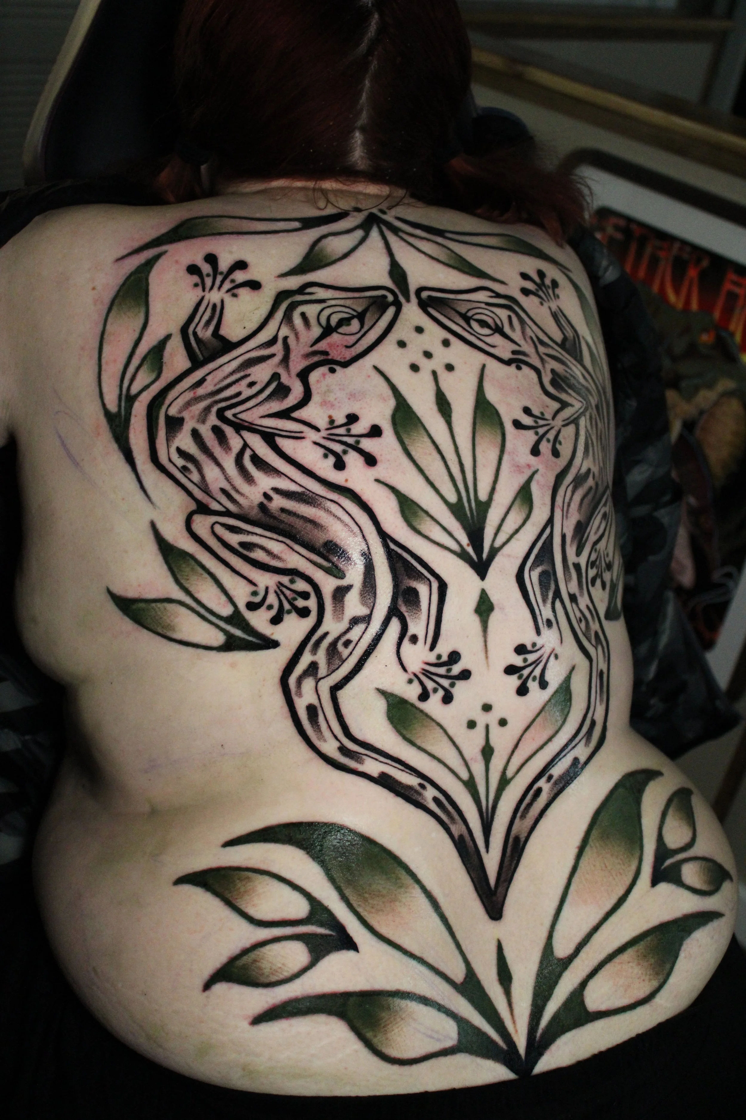 A tattoo on a person's back featuring a symmetrical floral and frog design in black ink with green accents.