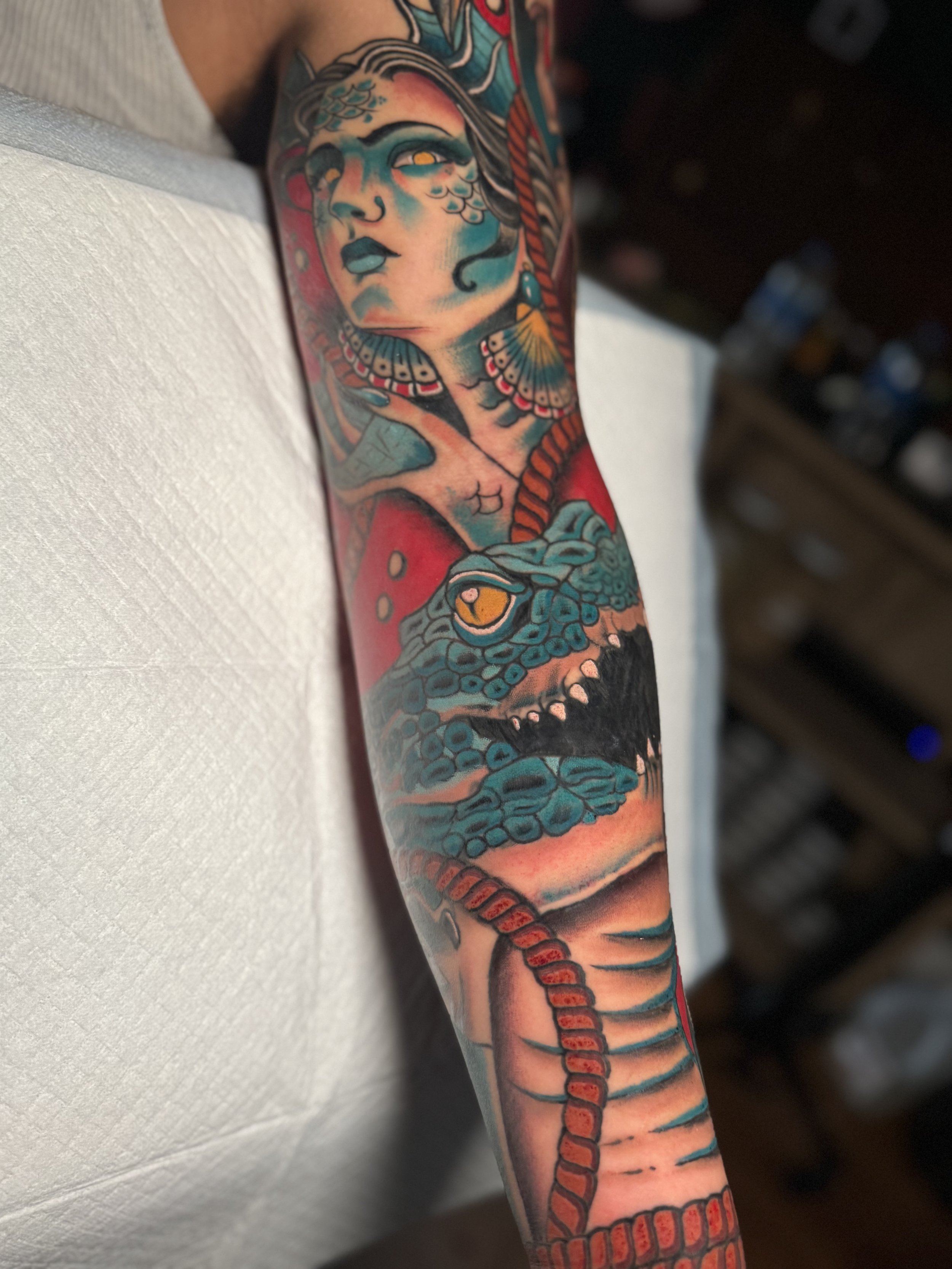 A colorful tattoo of a woman's face with blue lips, elaborate makeup, and floral and geometric designs on the arm, featuring a stylized snake intertwined with the design.