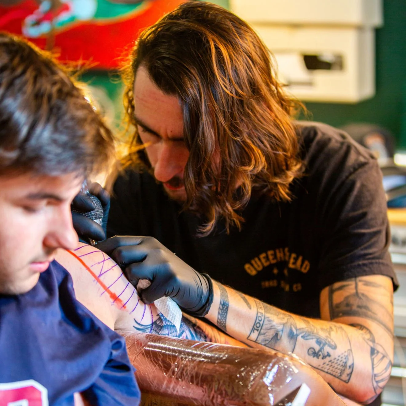 Tattoo artist with long hair and tattoos getting inked by a client in a tattoo studio.