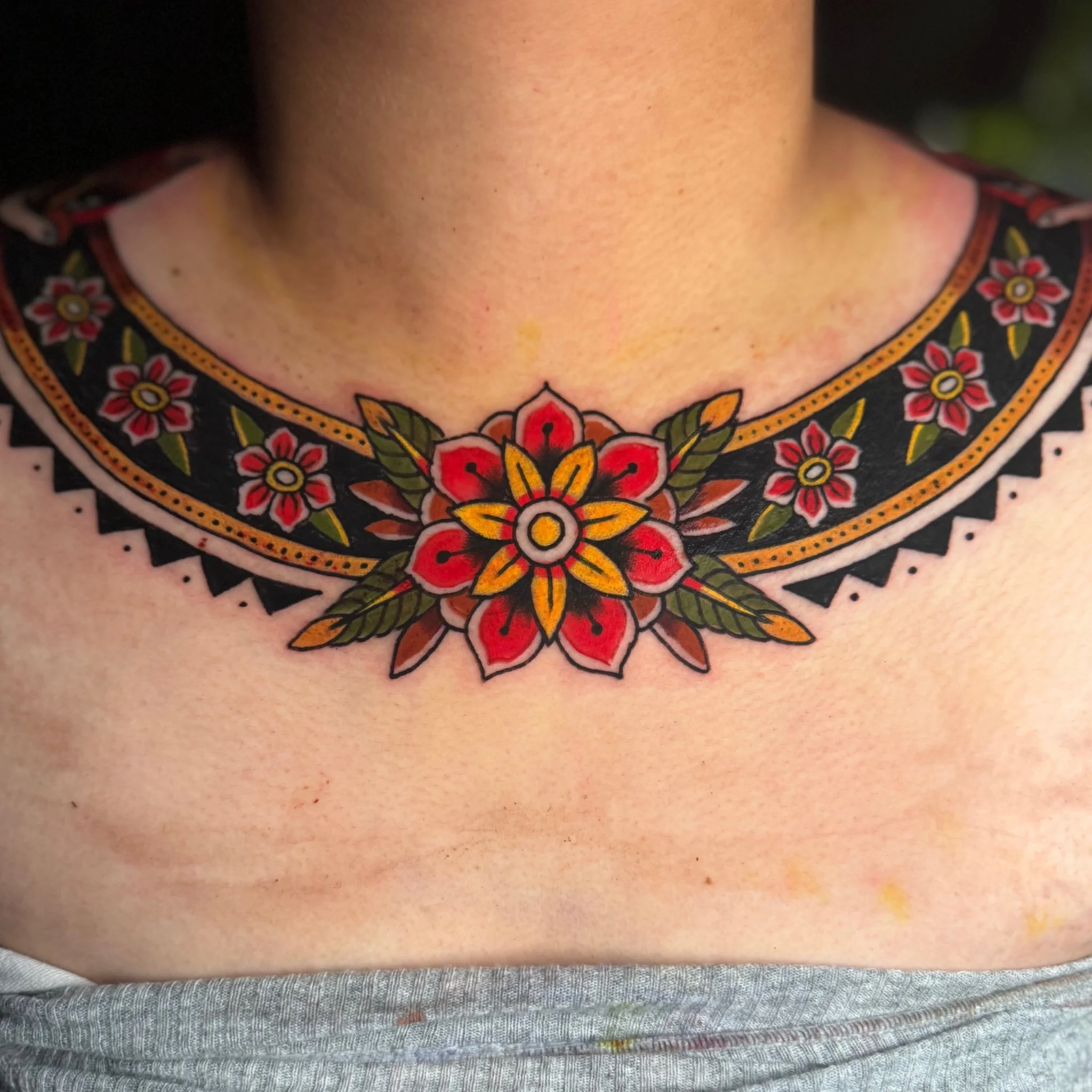 Close-up of a person's neck and upper chest, showing a vibrant, colorful, floral tattoo with red, orange, yellow, green, and black details on the skin.