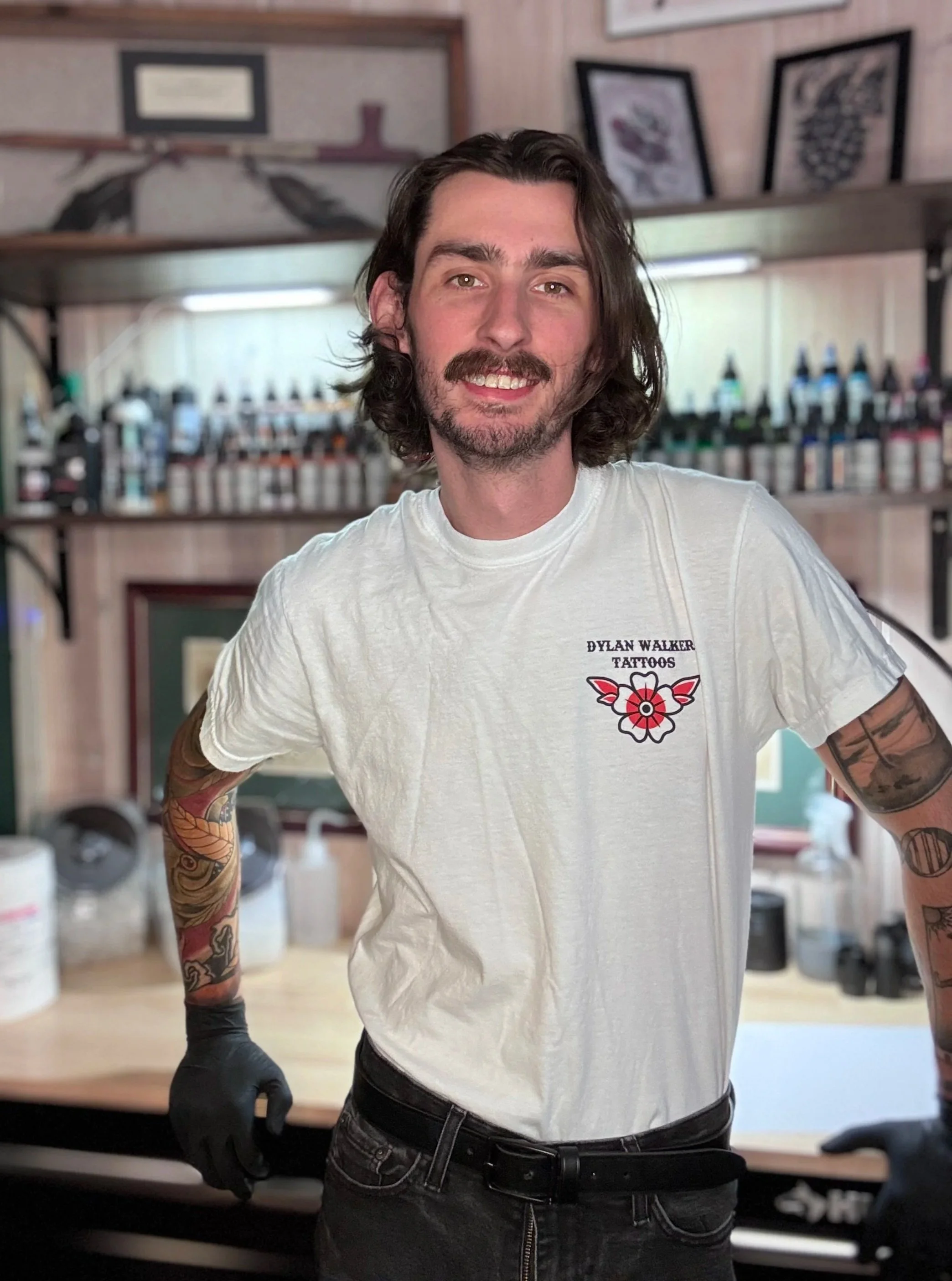 A man with long curly hair and tattoos on his arms, wearing a white T-shirt with a tattoo shop logo, standing in a tattoo studio with shelves of ink bottles behind him.