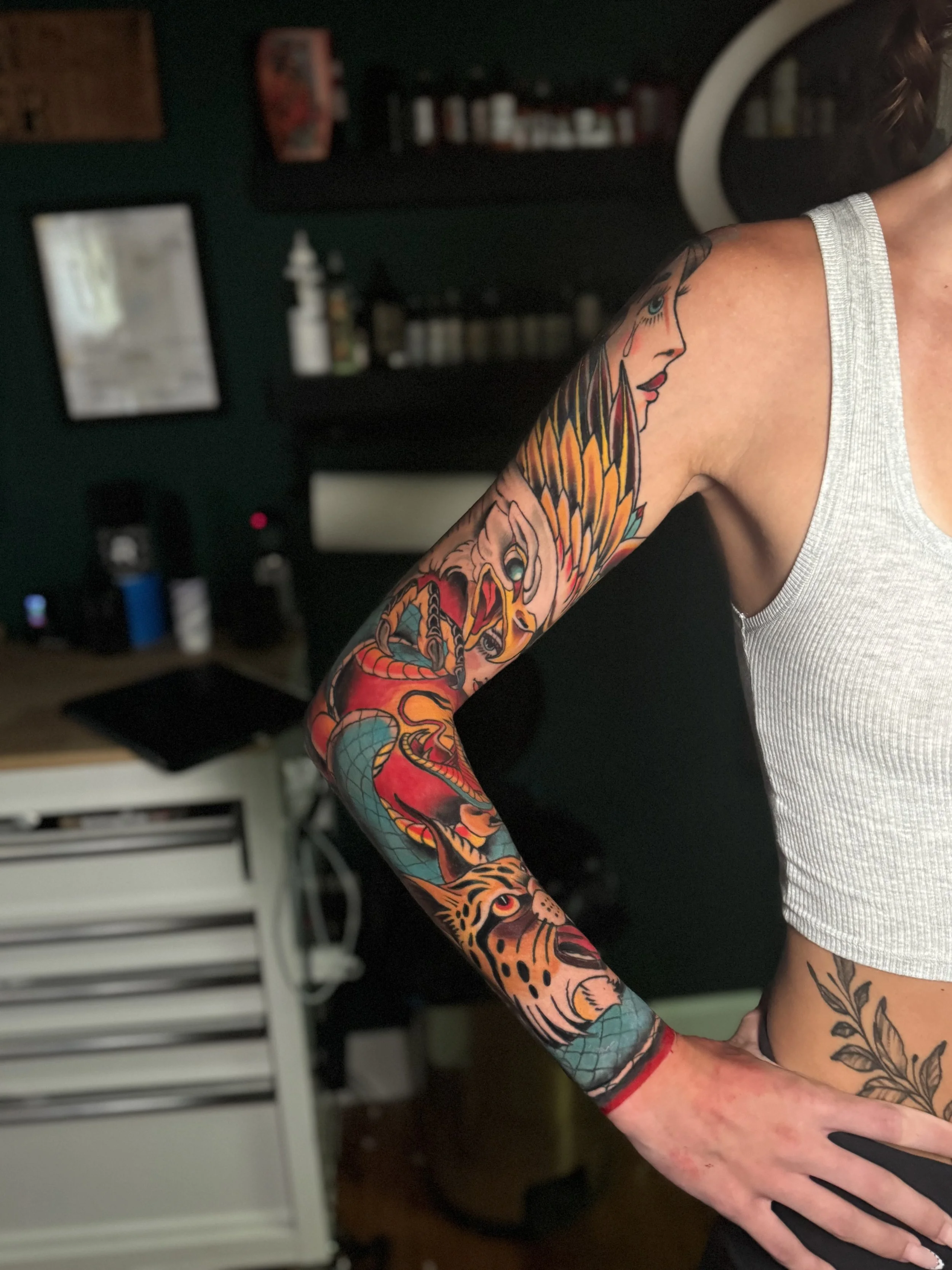 A person's arm with a colorful tattoo of a woman's face with flowing hair, a snake coiled around the arm, and a tiger's face near the wrist. The person is wearing a sleeveless gray shirt, and their hand is resting on their hip.