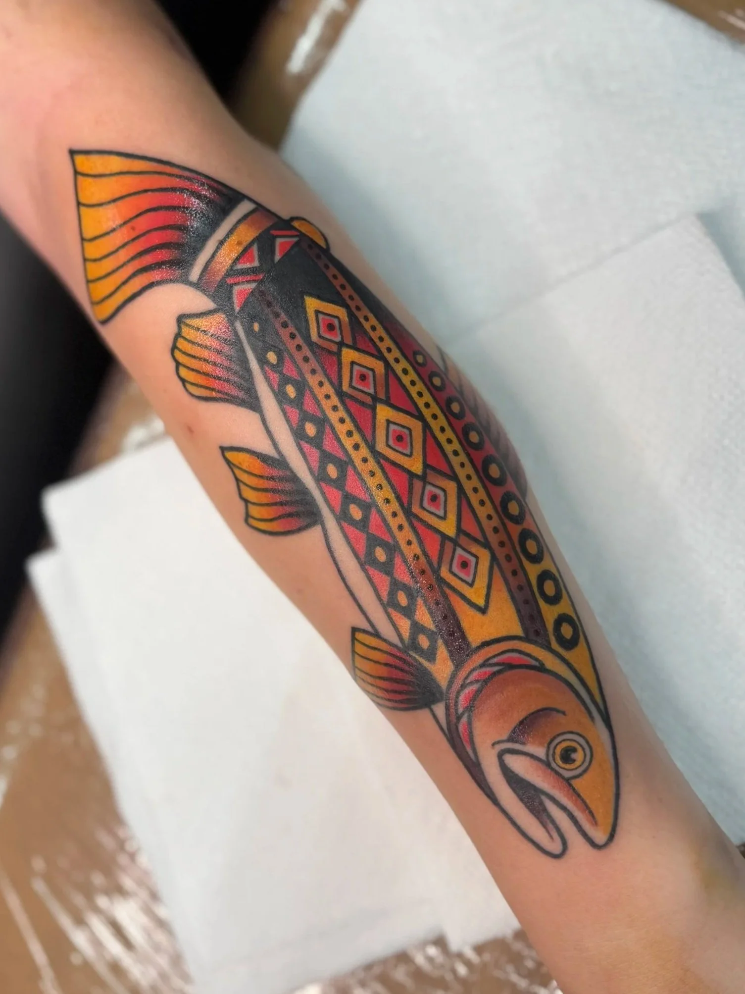 Colorful tattoo of a fish on person's arm, with orange, red, yellow, and black geometric and line patterns.