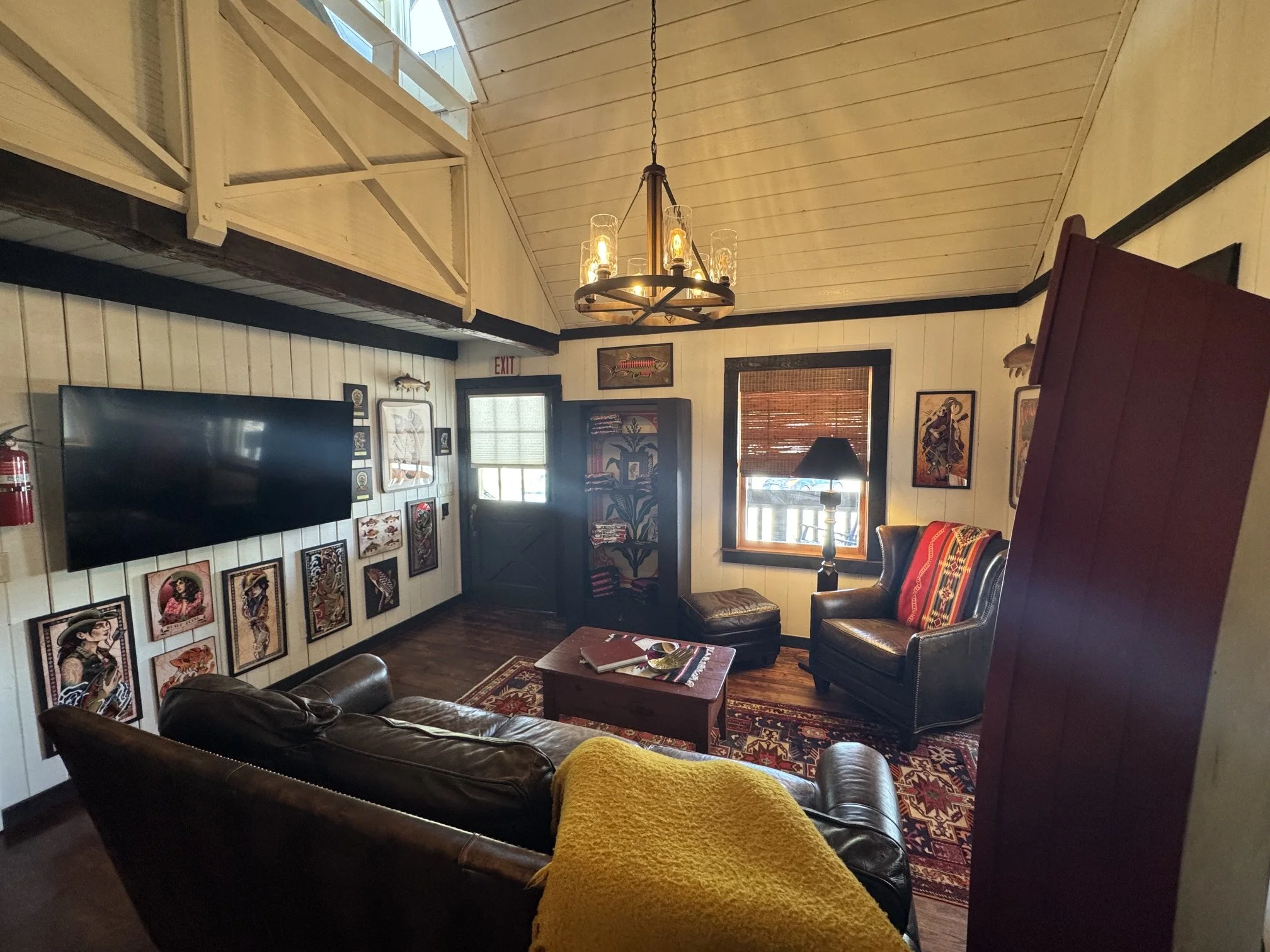 A cozy living room with a high, sloped ceiling and light-colored wooden walls. There is a chandelier hanging from the ceiling with exposed bulbs. The room has various framed artwork on the walls, including a large fish picture. Furniture includes a l