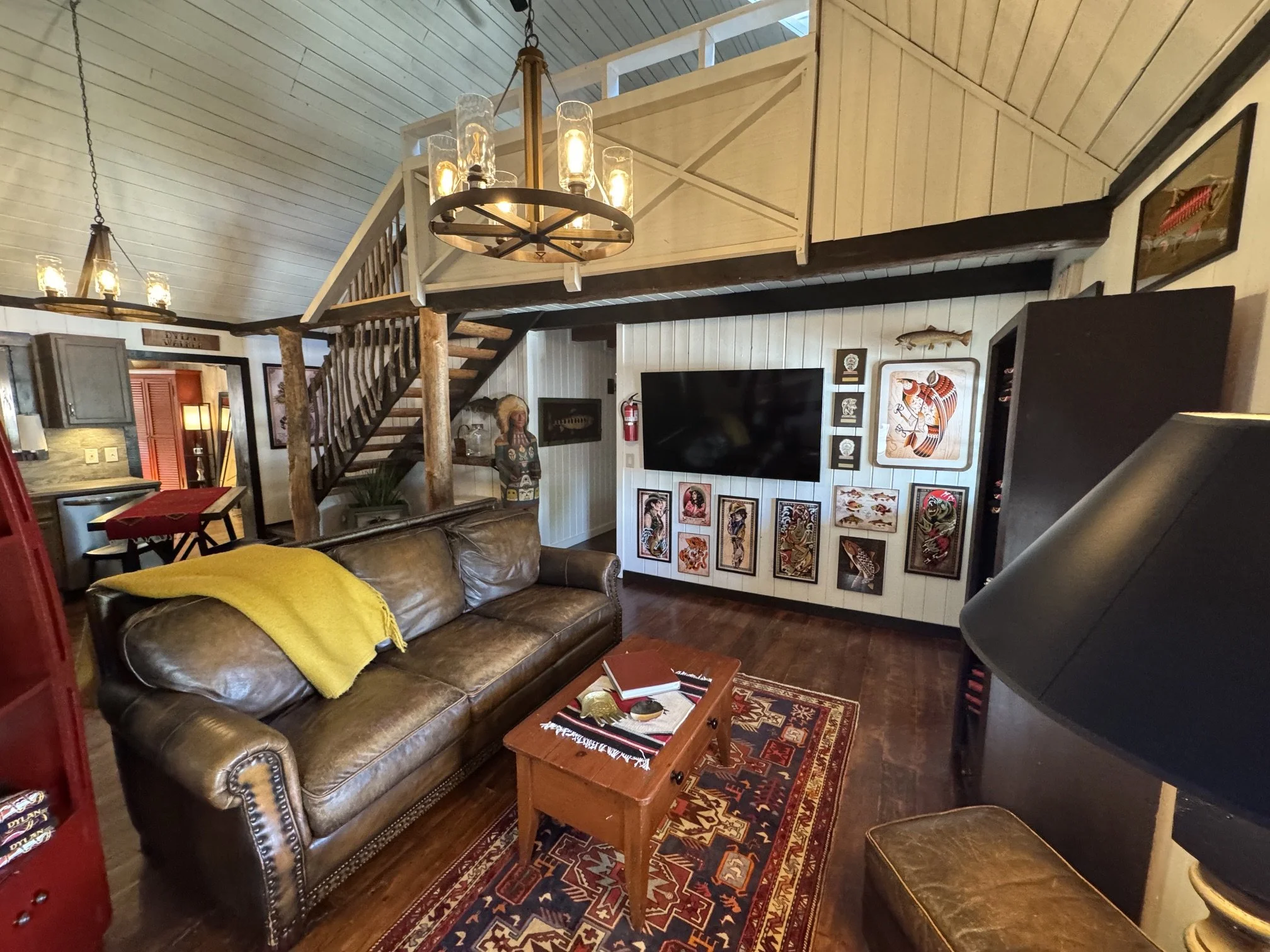 A cozy living room with a brown leather sofa, a yellow blanket, a wooden coffee table, and a patterned rug. A large flat-screen TV is mounted on a white paneled wall decorated with framed artwork. A rustic chandelier hangs from the ceiling, and a sta