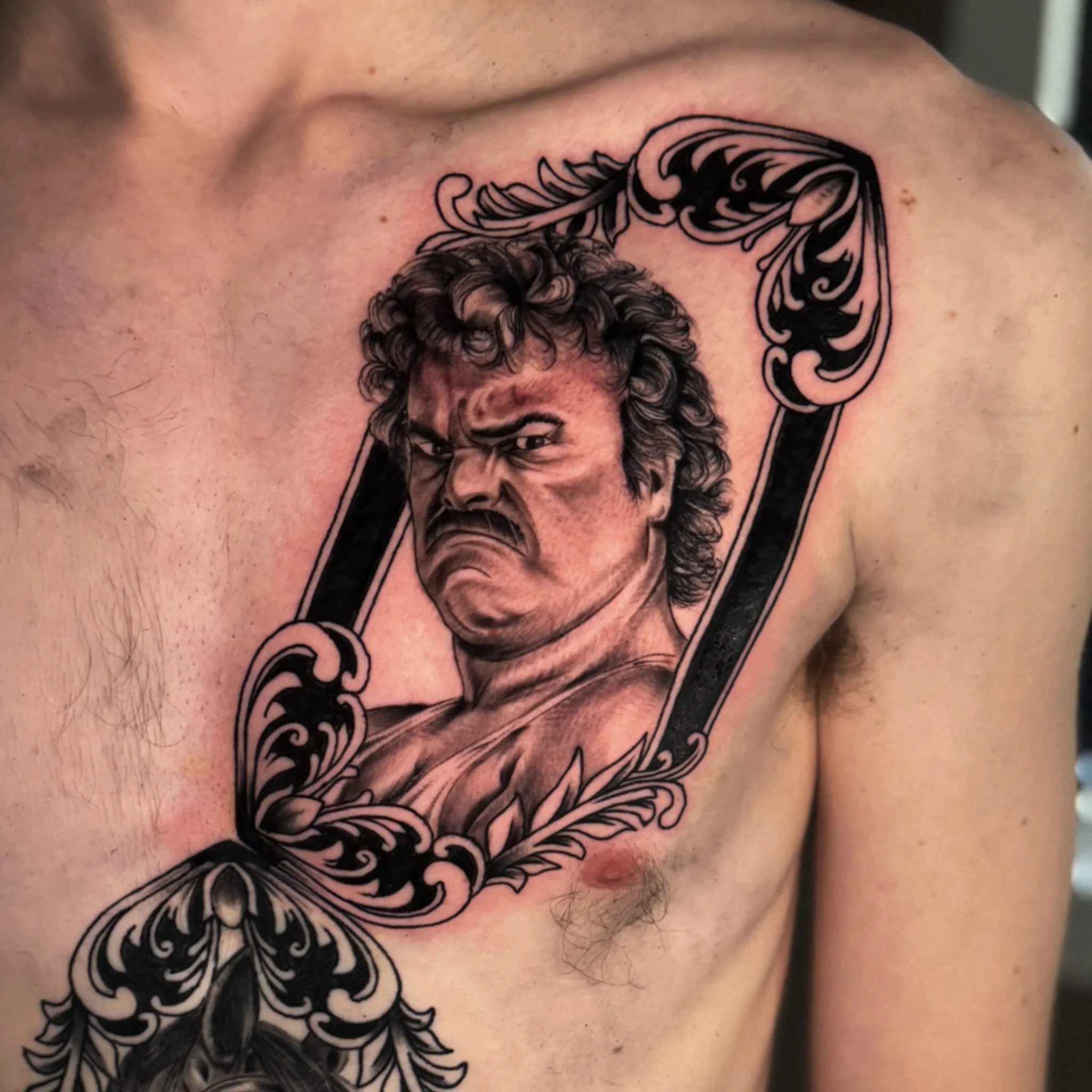 A tattoo of a person's face with curly hair and an intense expression on their forehead, surrounded by decorative black and gray linework on the shoulder.