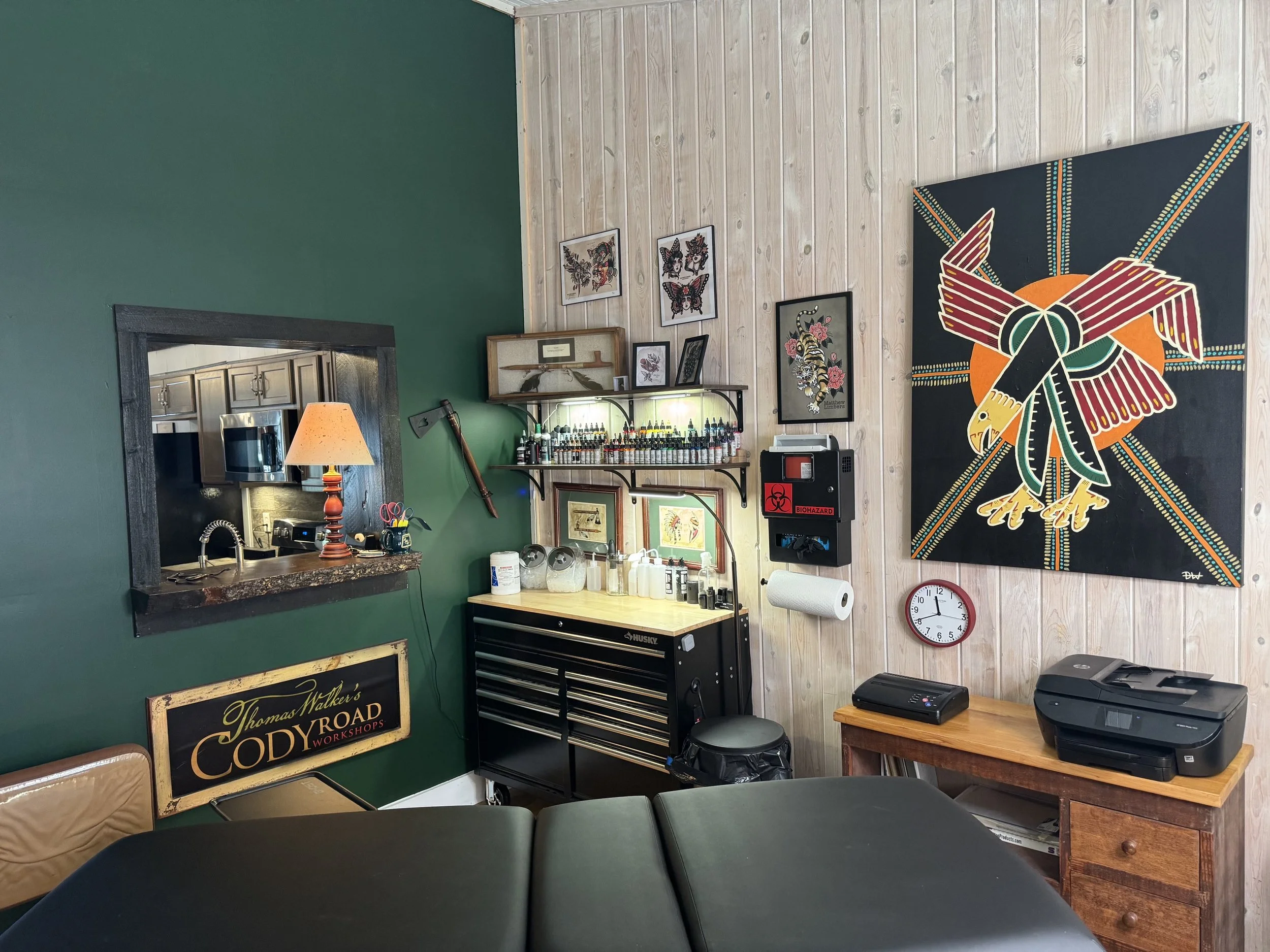 Interior of a tattoo studio with framed artwork on the wall, a workbench with craft supplies, a small table with a printer and a paper roll, and a large colorful graffiti-style eagle mural on the wall.