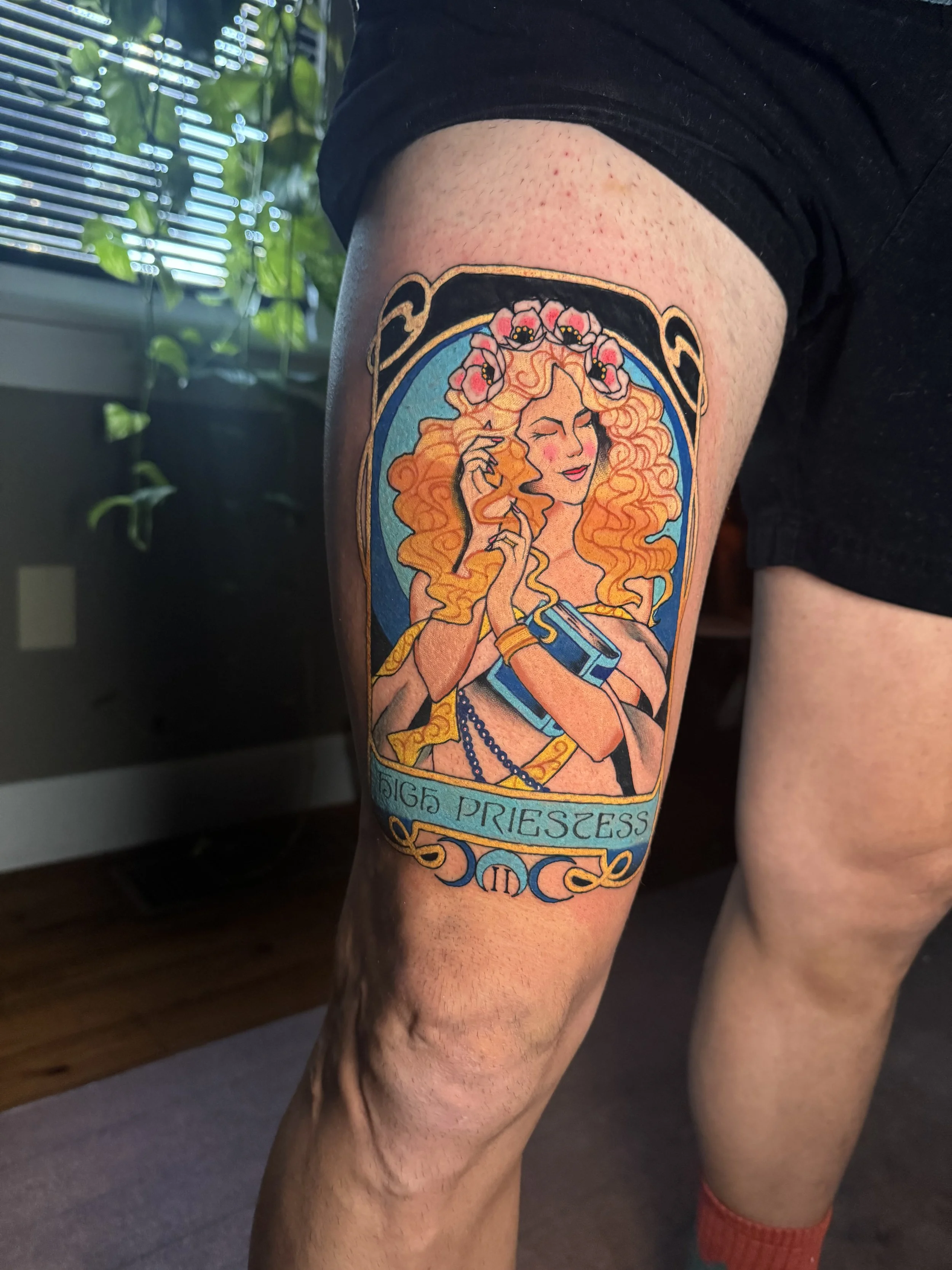 Tattoo of a woman with long curly hair, holding a scroll, with flowers in her hair, on a person's thigh.