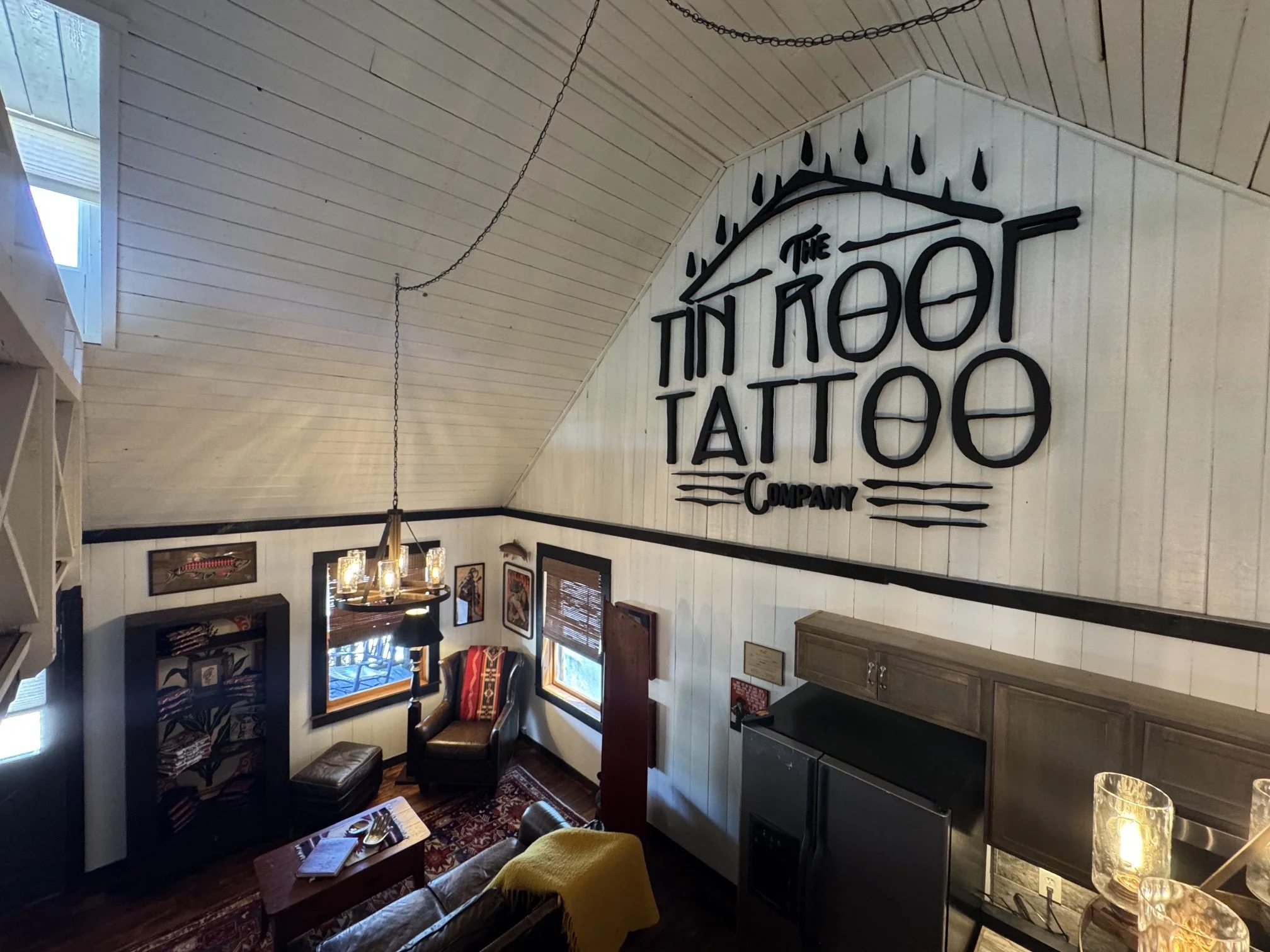 Interior of a cozy restaurant or café with white paneled walls, wooden furniture, and artwork. There is a large sign on the wall that reads 'The Tin Roof Tattoo Company' with a stylized roof design. The room has a high, angled ceiling with chains han