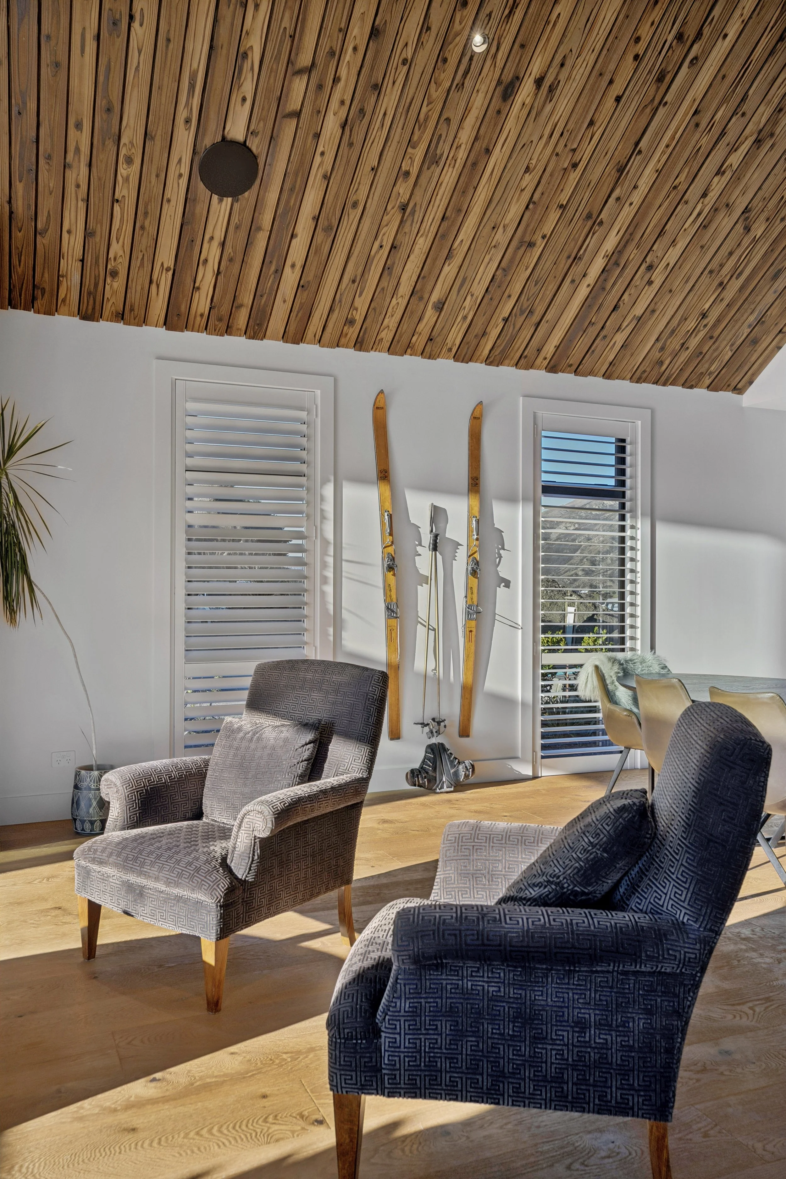 A cozy living room with two patterned armchairs, a pair of vintage skis mounted on the white wall, and a window with white shutters letting in natural light. The room features a wooden ceiling and hardwood flooring.