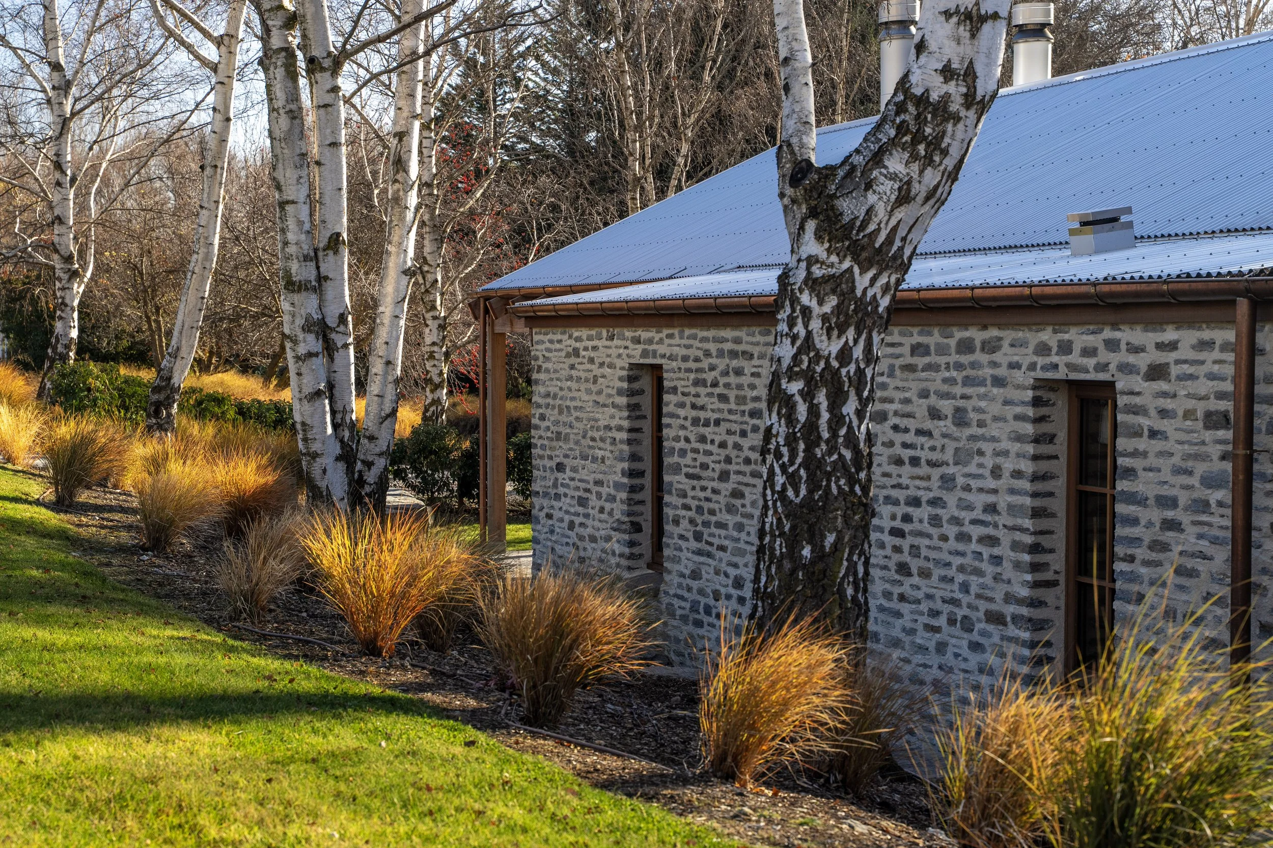 A stone house with a blue metal roof surrounded by trees and ornamental grass in fall or winter.