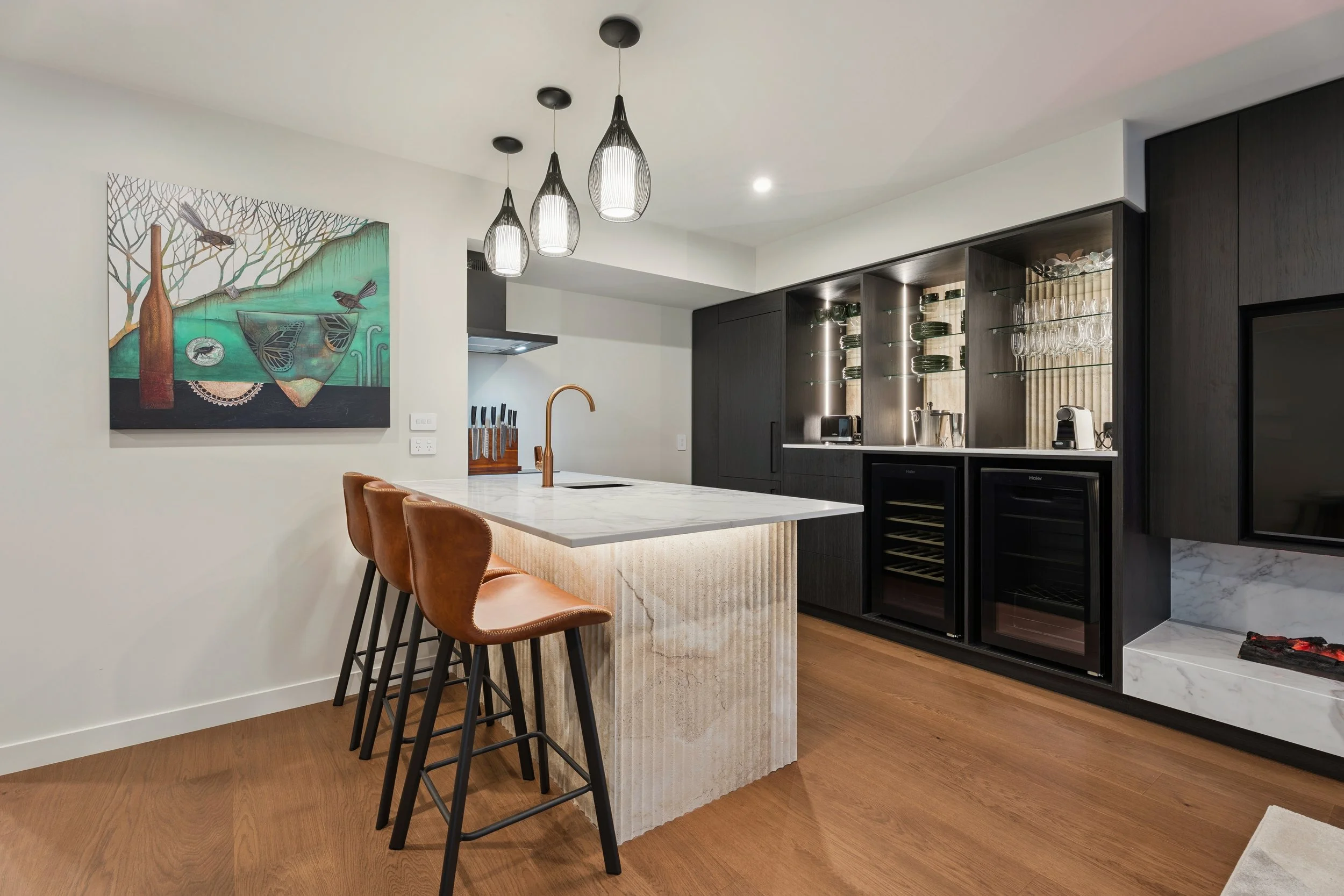 Modern kitchen with a marble island, brown bar stools, black cabinetry, geometric pendant lights, and artwork on the wall.