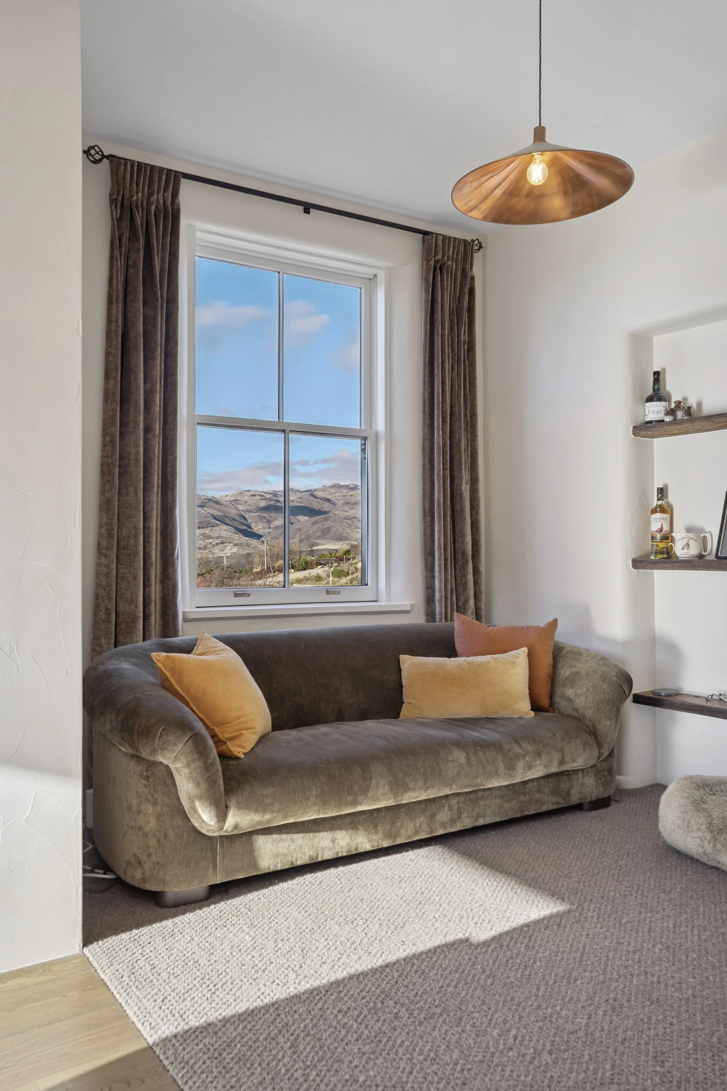 A living room with a gray velvet couch and orange pillows, a large window with brown curtains showing a mountain landscape, a wooden ceiling lamp, shelves with bottles and decorative items, and a mix of beige and brown rugs.
