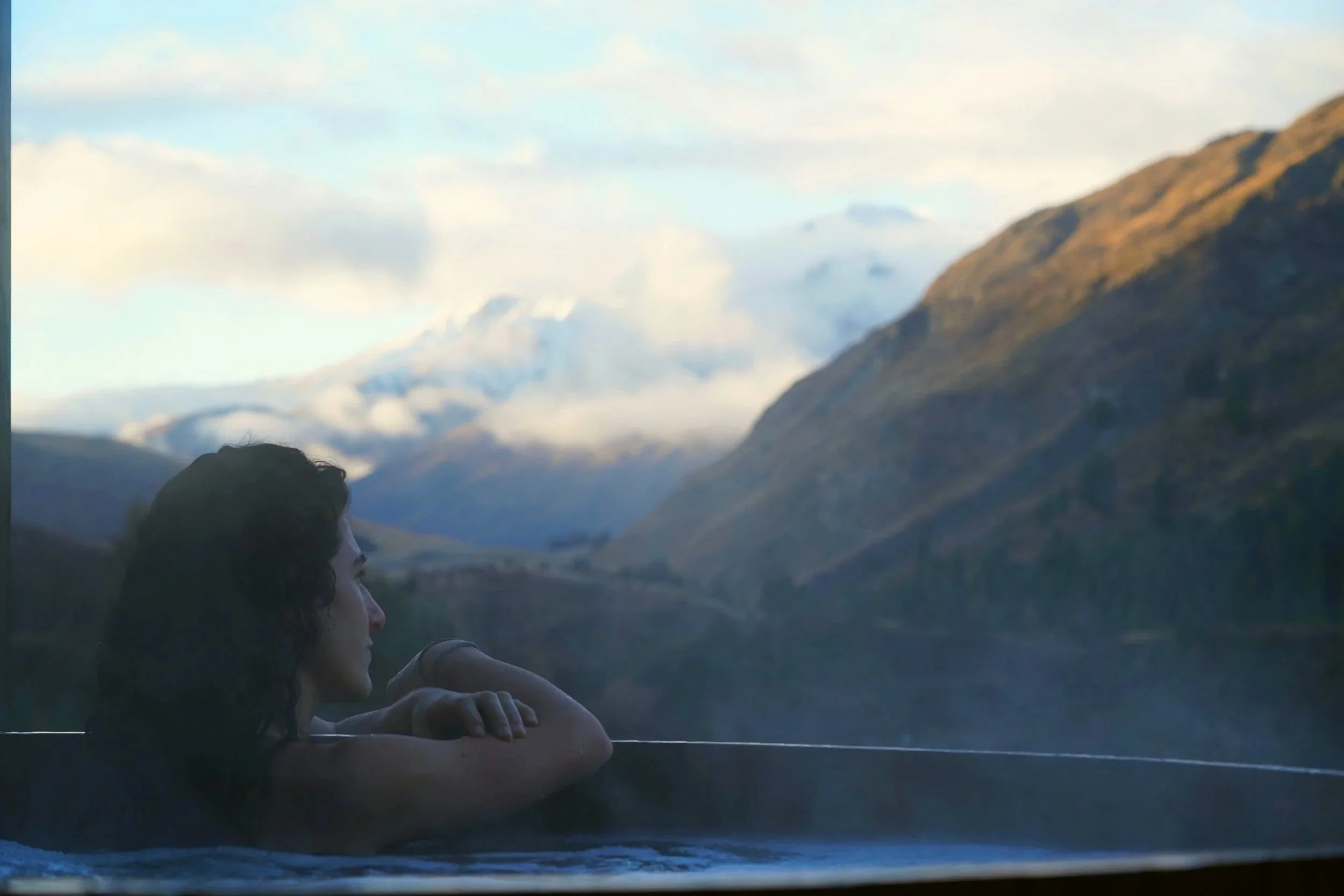 A person with dark, curly hair is resting their arms on a railing, looking at a scenic mountain landscape with snow-capped peaks and cloudy sky in the background.