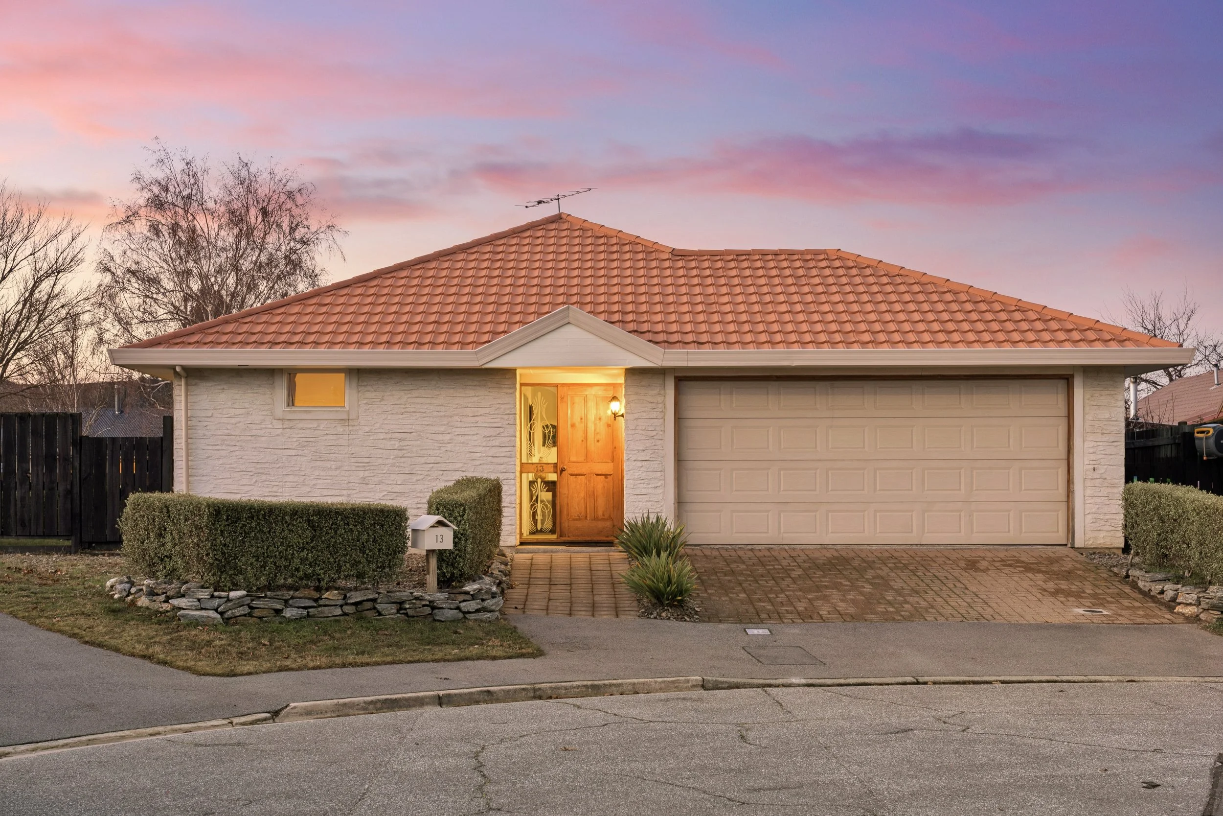 A house with a tiled roof, lit-up front door, and a garage, surrounded by well-maintained bushes and trees under a colorful sky at sunset.