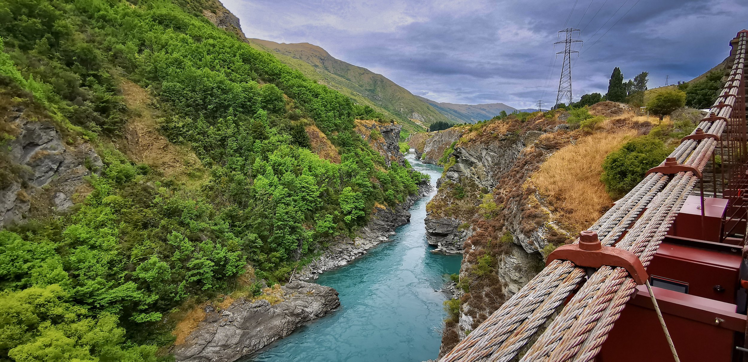 View of a river running through a canyon with green trees on the left and dry grass and shrubs on the right, seen from a bridge with a red railing, in a mountainous area with power lines and cloudy sky.
