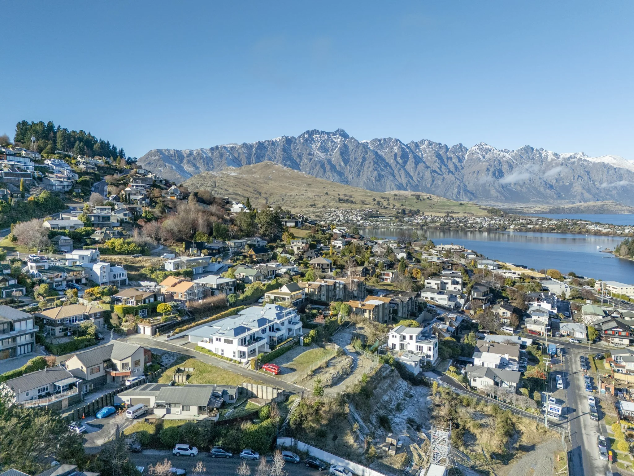 A scenic landscape of a residential area by a body of water with mountains in the background under a clear blue sky.