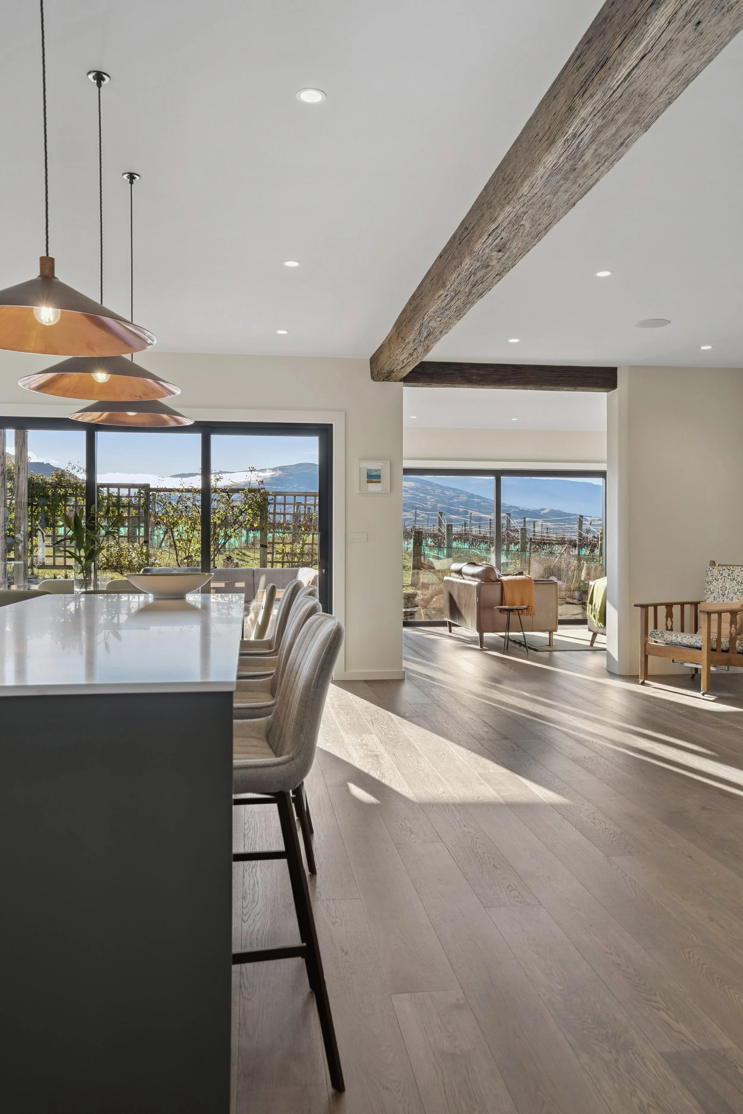 Modern dining room and living room with large windows showing a mountain landscape, exposed wooden beams, a dining table with upholstered chairs, and a seating area with armchairs and a view of the outdoors.