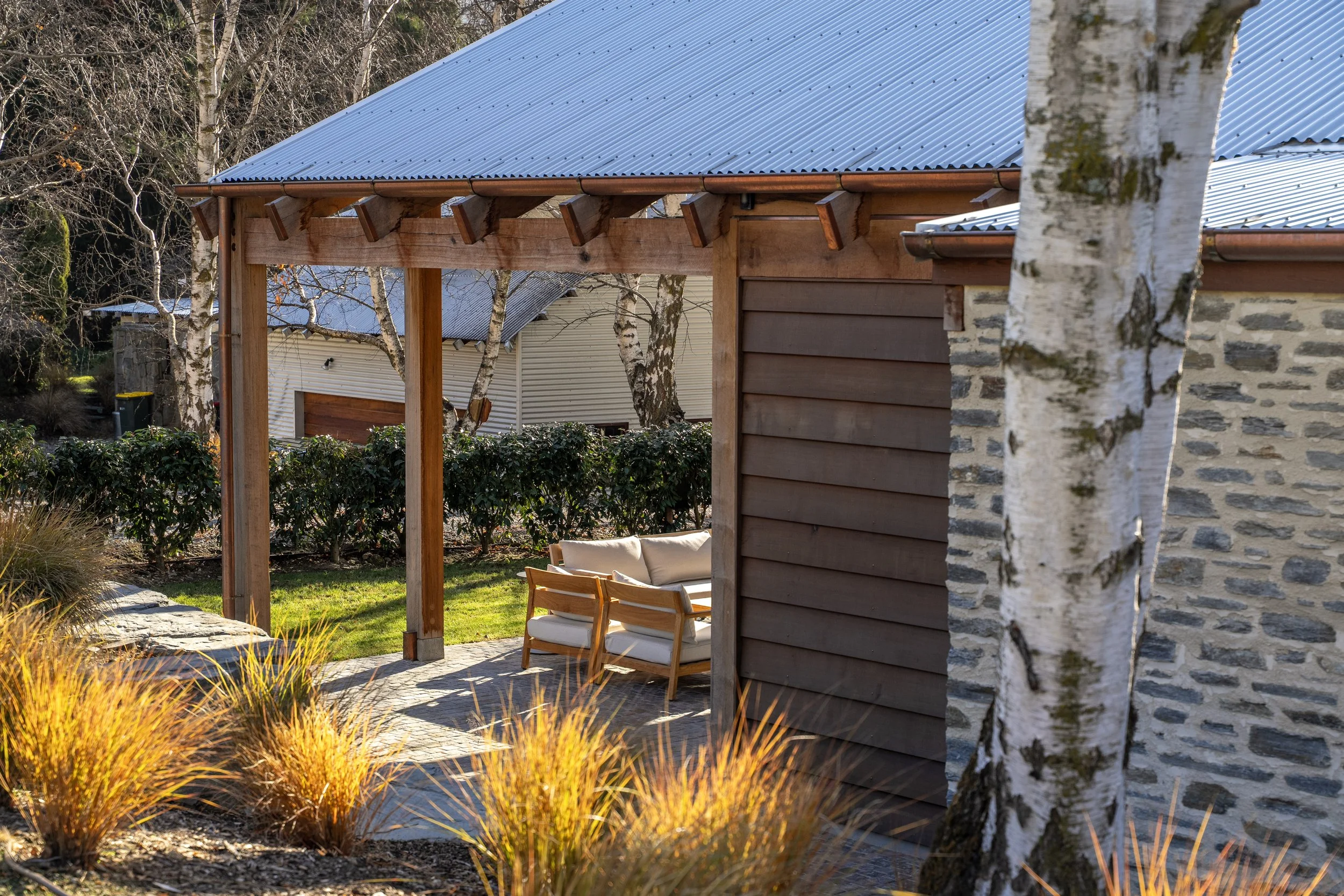 Outdoor patio with a wooden pergola, seating area with beige cushions, surrounded by decorative grasses and trees, with neighboring buildings in the background.