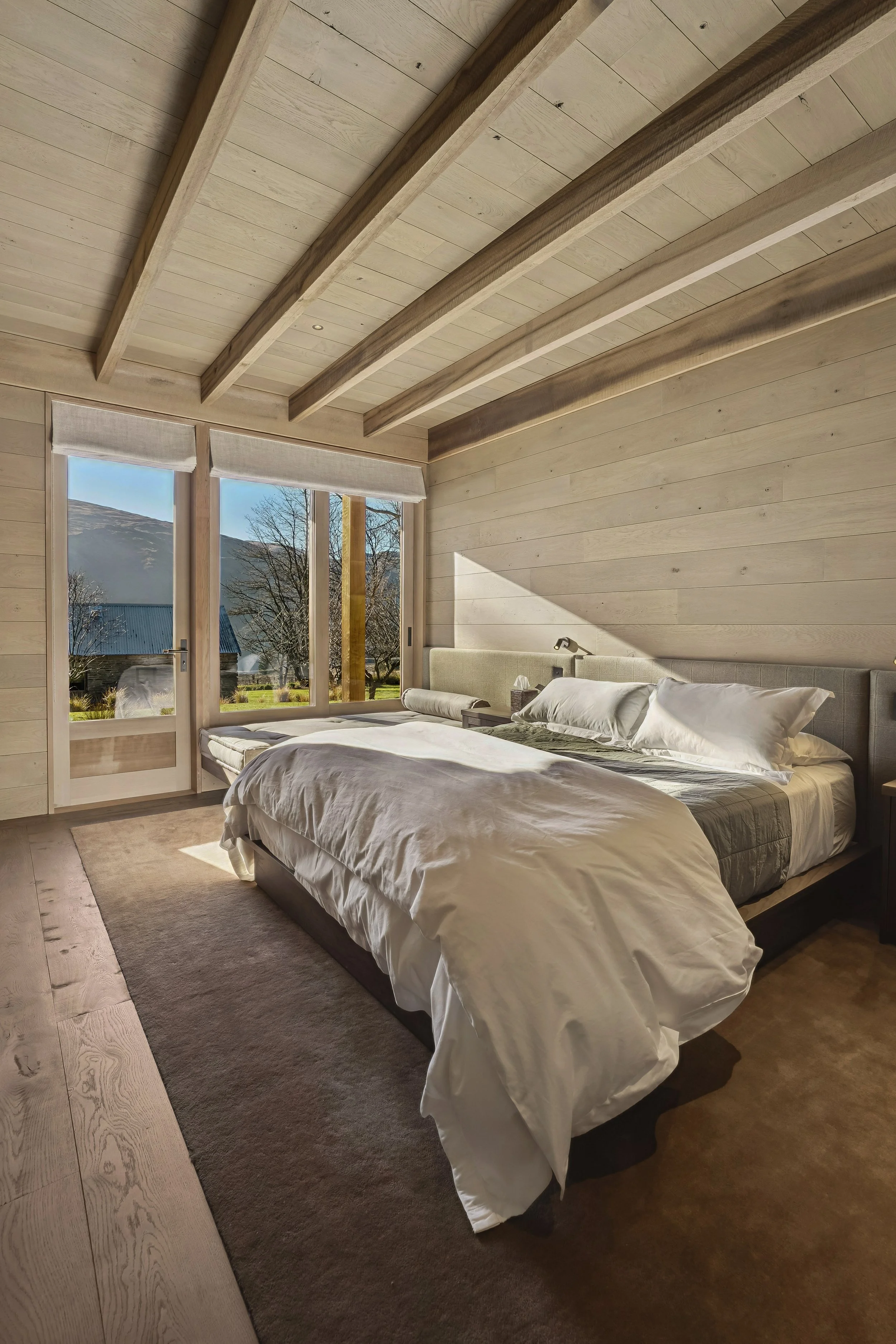 A bedroom with a large bed covered in white bedding and pillows, wooden walls and ceiling, large windows showing an outdoor landscape, and a beige rug on the wooden floor.