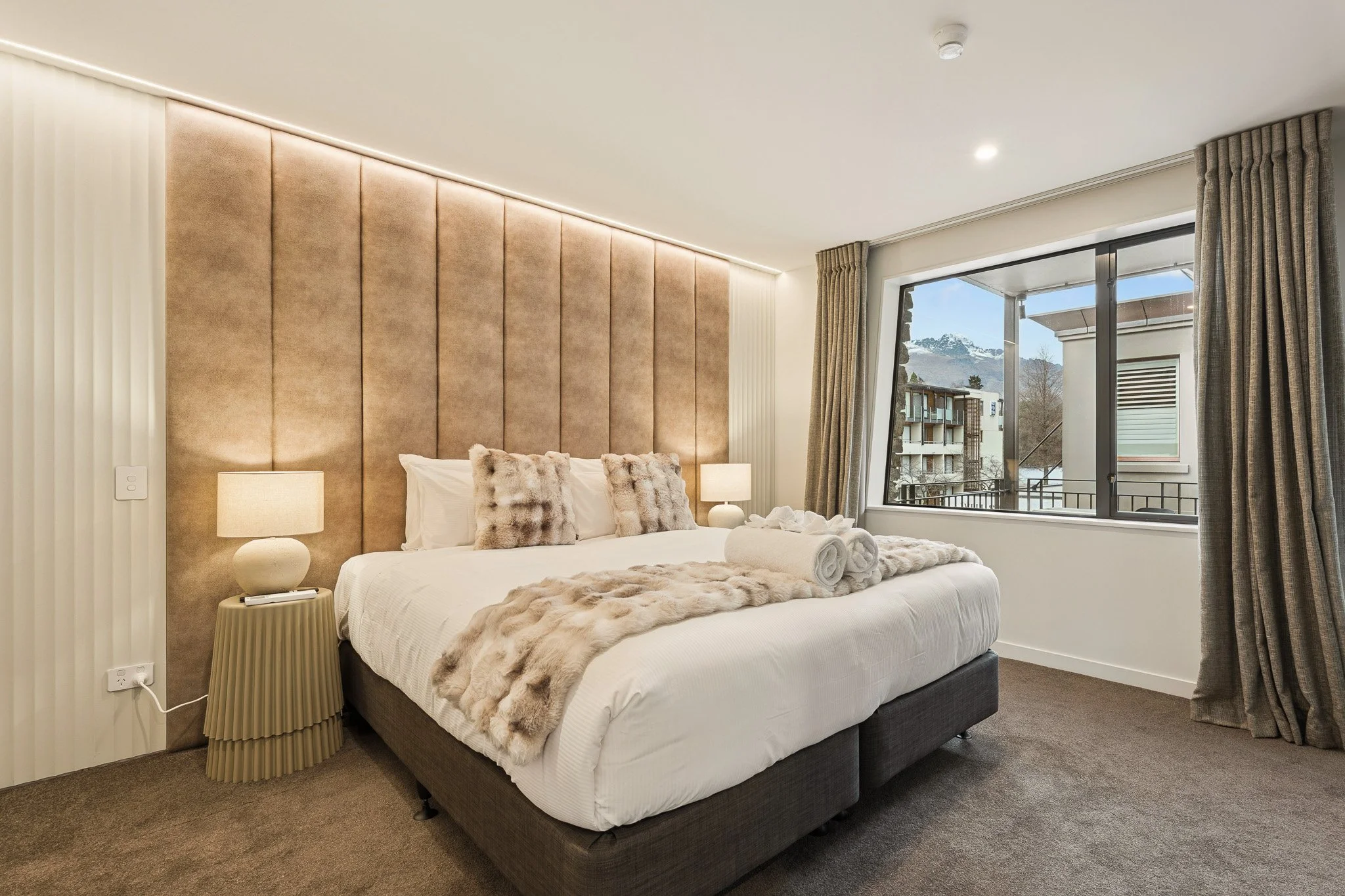 Modern bedroom with a large bed, beige upholstered headboard, fur throw pillows, matching fur throw blanket, bedside lamps, window with beige curtains, mountain view outside.